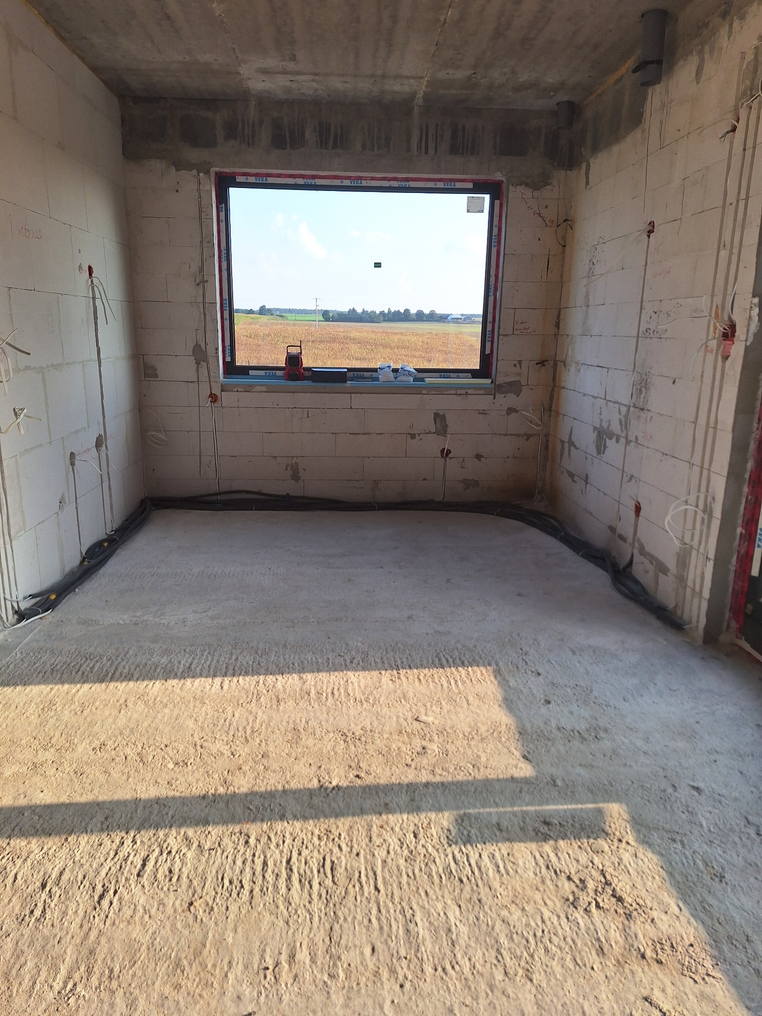 Surowy pokój z oknem na pole. Widoczne kable elektryczne w ścianach i rury ogrzewania podłogowego przed wylewką. Stan deweloperski, ściany z betonu komórkowego.