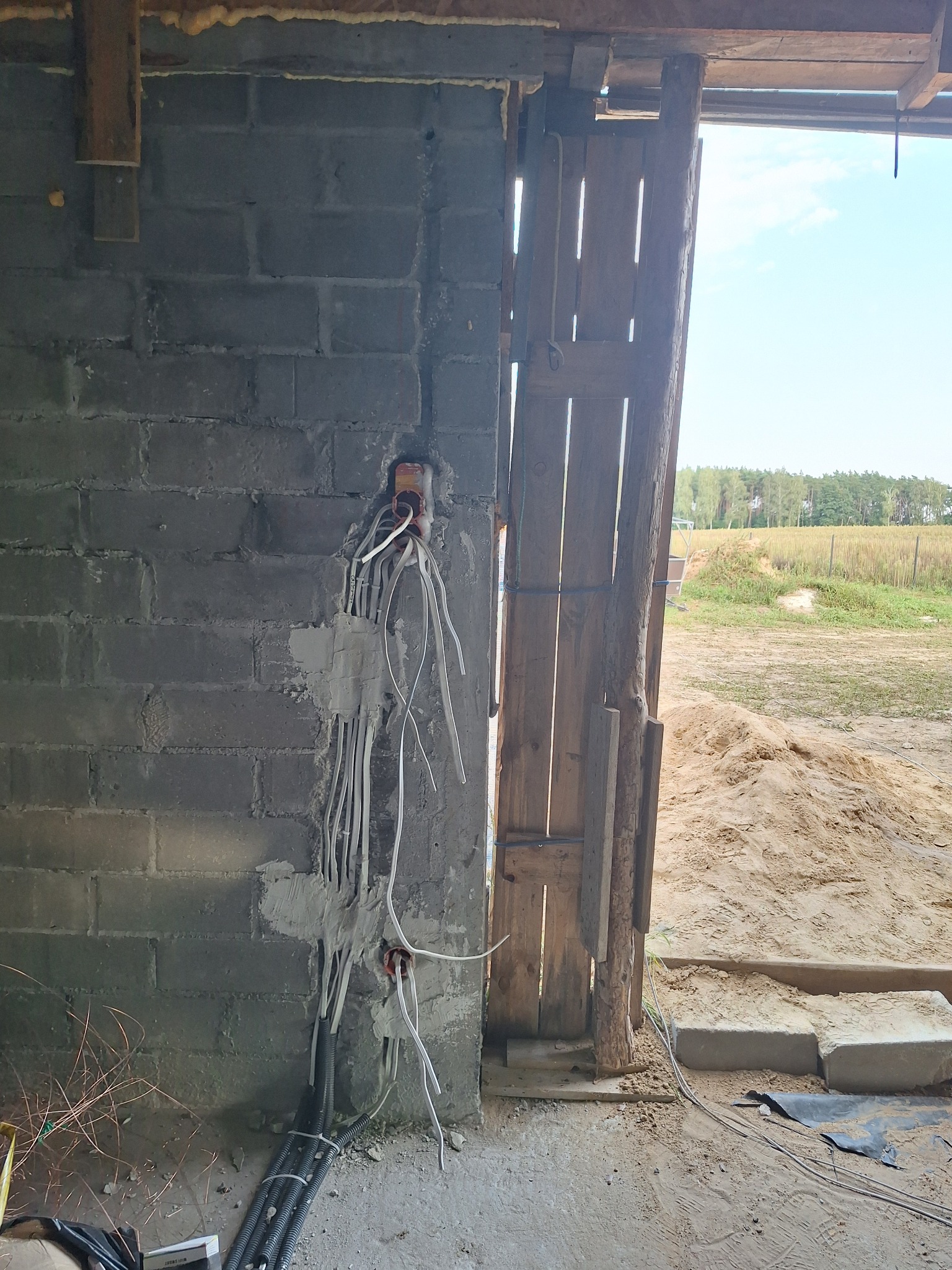 Surowa ściana z szarego betonu komórkowego z przewodami elektrycznymi wychodzącymi z puszek, widok na zewnątrz przez drewniane drzwi.