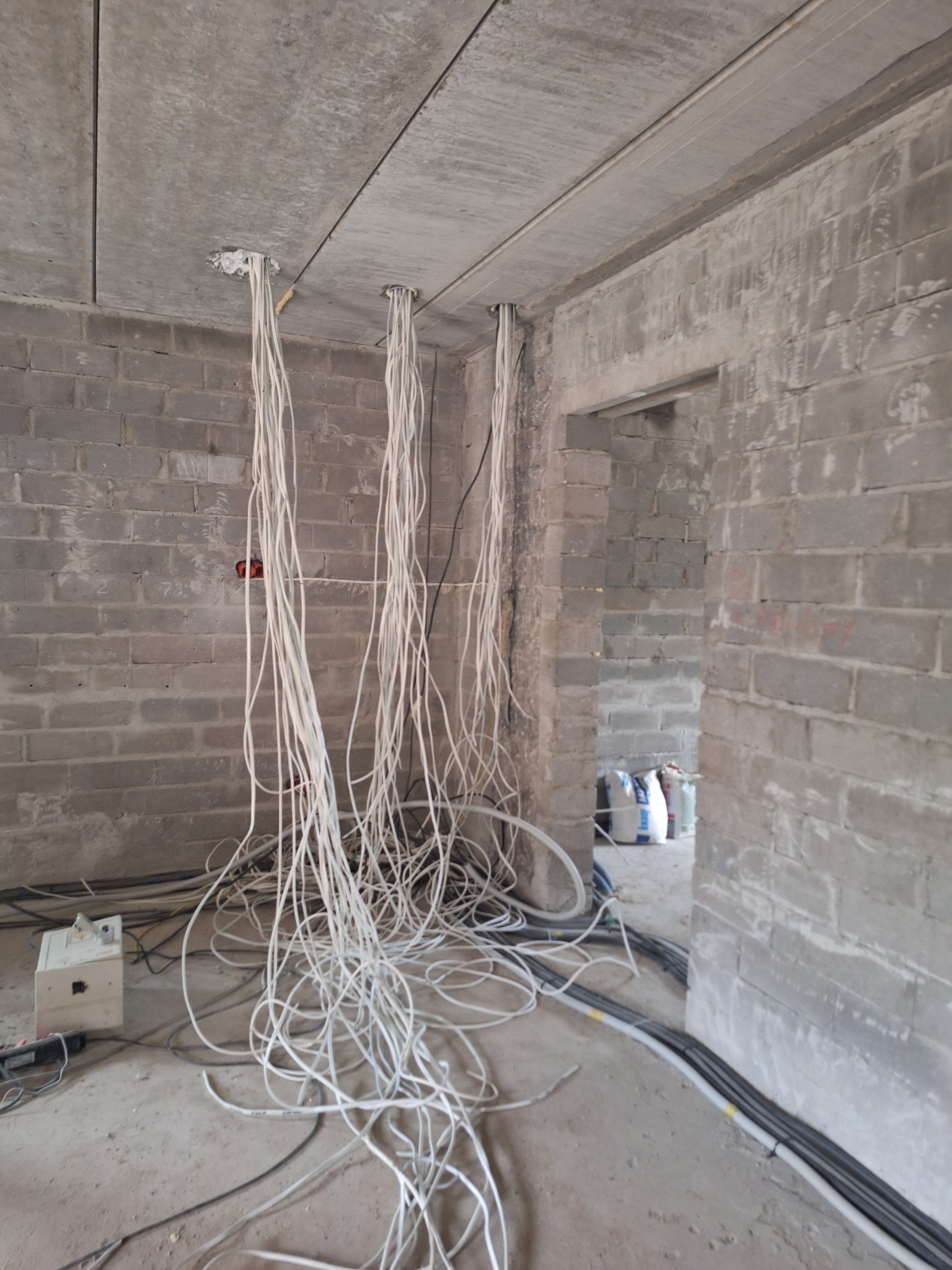 Surowy stan: kable zwisają z sufitu w szarym, ceglanym wnętrzu w trakcie instalacji elektrycznej. Widoczne przewody na podłodze i surowy betonowy sufit.