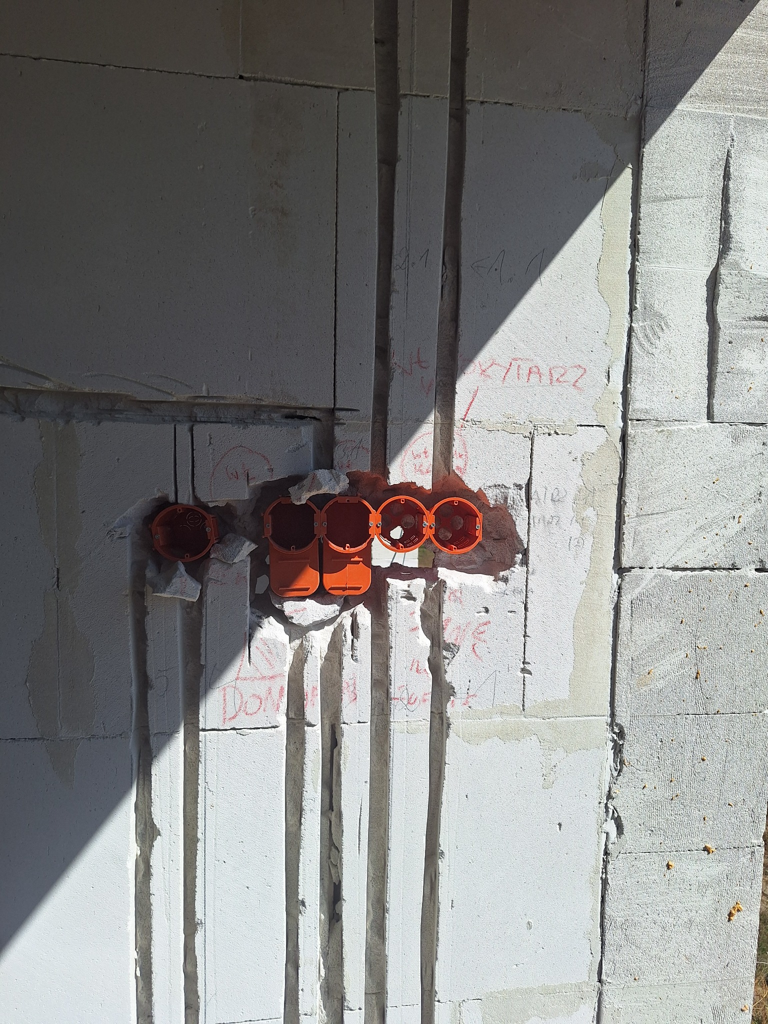 Biała ściana z betonu komórkowego z wyżłobionymi bruzdami i pomarańczowymi puszkami elektrycznymi. Widoczne napisy na ścianie i cień. Instalacja elektryczna w trakcie realizacji.
