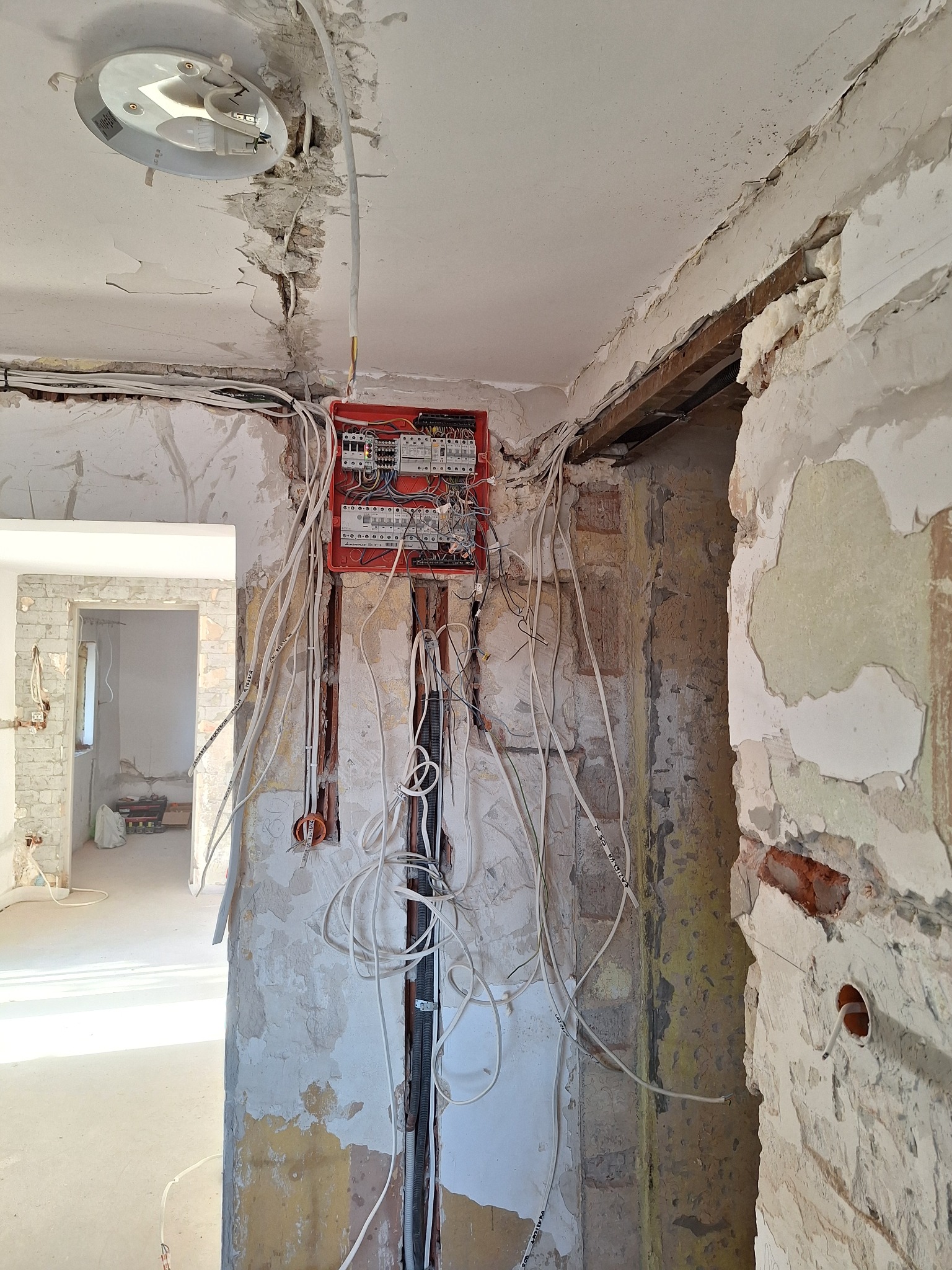 Rozdzielnia elektryczna z licznymi przewodami w surowym wnętrzu podczas remontu. Widoczne ślady po kuciu w ścianach i suficie, odsłonięta instalacja elektryczna.