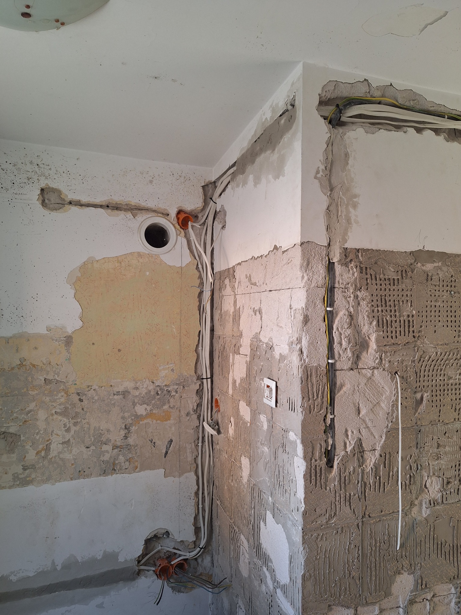 Ściana w trakcie remontu z widocznymi przewodami elektrycznymi w bruzdach, przygotowana pod tynkowanie. Fragment ściany z pozostałościami po płytkach.