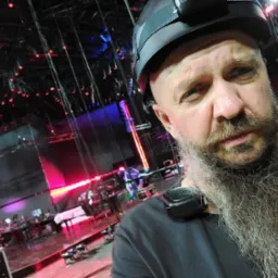 Autoportret brodatego mężczyzny w kasku z lampką czołową na tle konstrukcji oświetleniowej sceny koncertowej z czerwonym i niebieskim światłem.