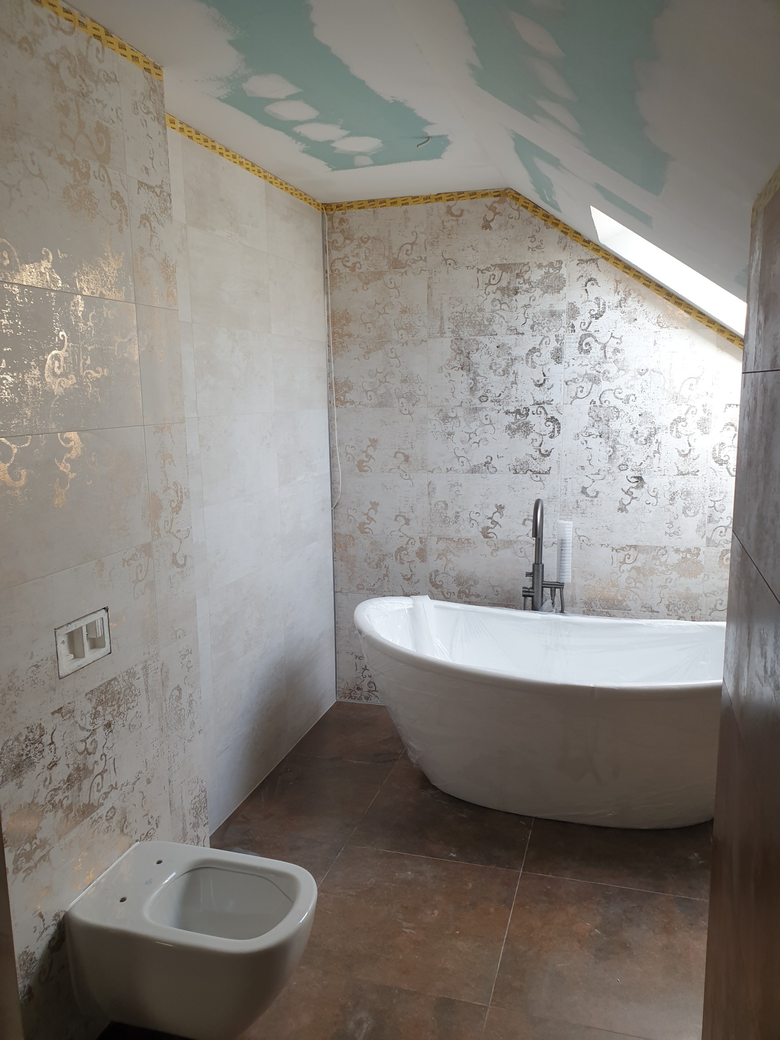 Remontowana łazienka w Sosnowcu: biała wanna owalna zabezpieczona folią, podwieszana toaleta oraz ściany wyłożone płytkami z delikatnym wzorem, sufit częściowo zagruntowany.