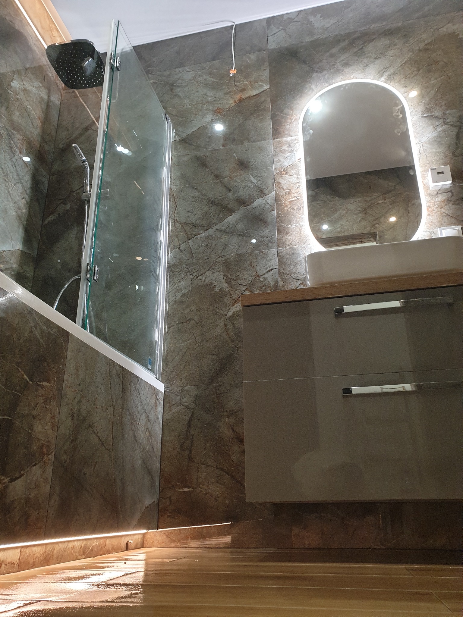 Nowoczesna łazienka z marmurowymi płytkami, kabiną prysznicową ze szklanym panelem, podświetlanym lustrem nad umywalką w szafce z szarymi frontami i oświetleniem LED przy podłodze.