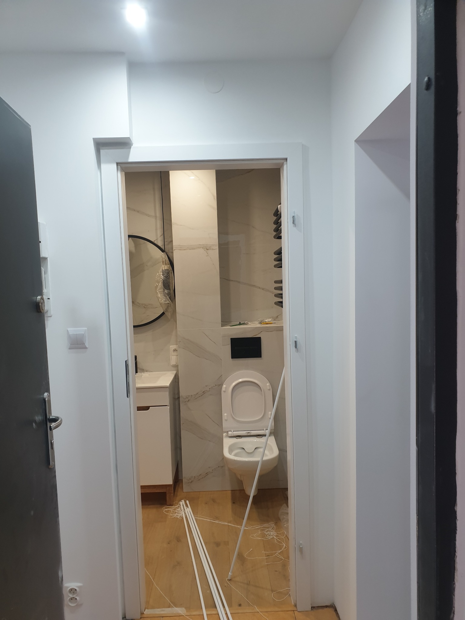 Widok na wnętrze łazienki z białymi marmurkowymi płytkami, umywalką z szafką i toaletą, widziany z korytarza przez odnowione ościeżnice drzwiowe, z widocznymi listwami przypodłogowymi na podłodze.