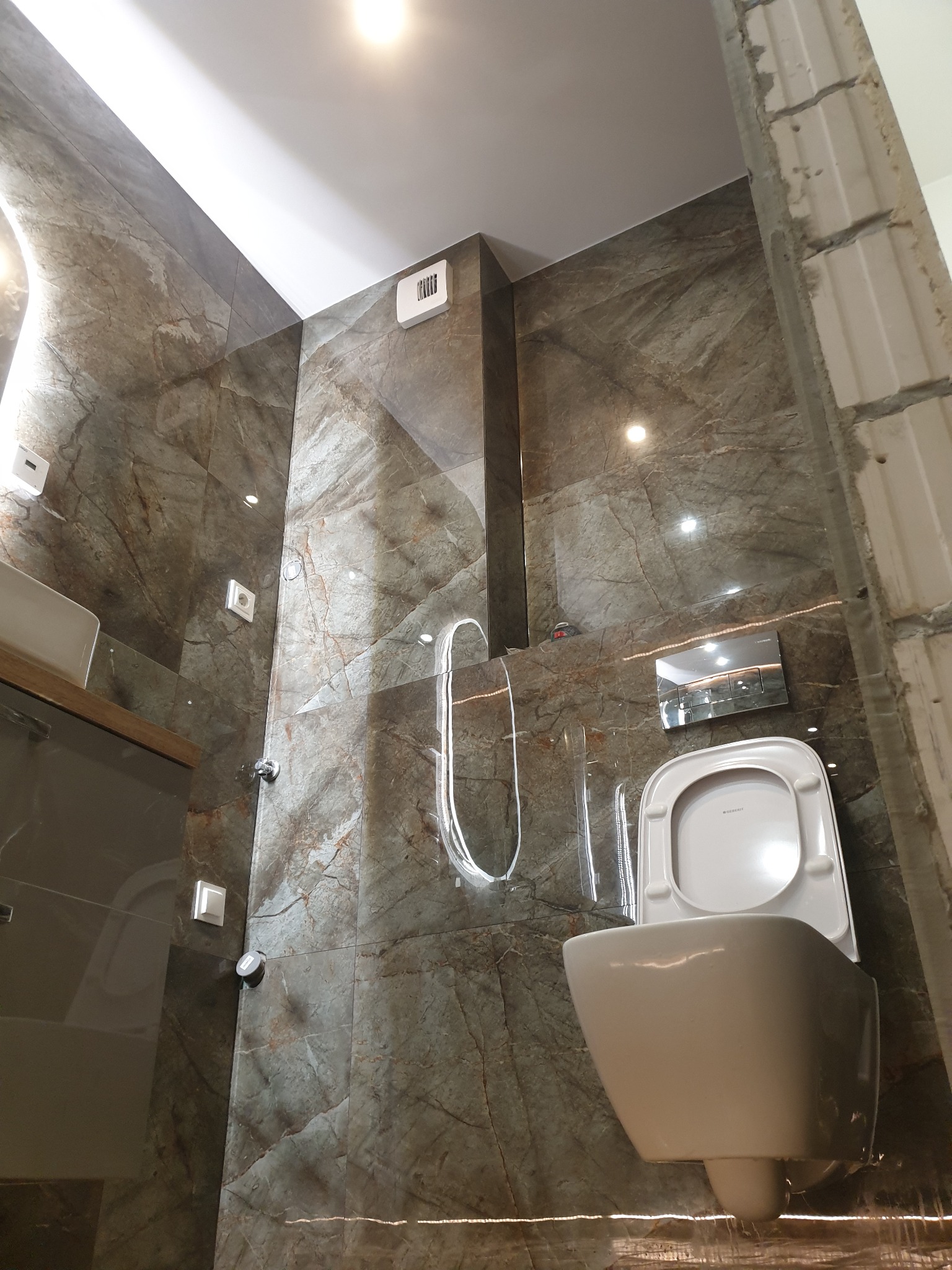 Wisząca toaleta z deską Geberit na tle ściany wyłożonej brązowymi płytkami imitującymi marmur, widoczny fragment ściany w stanie surowym, po prawej stronie.