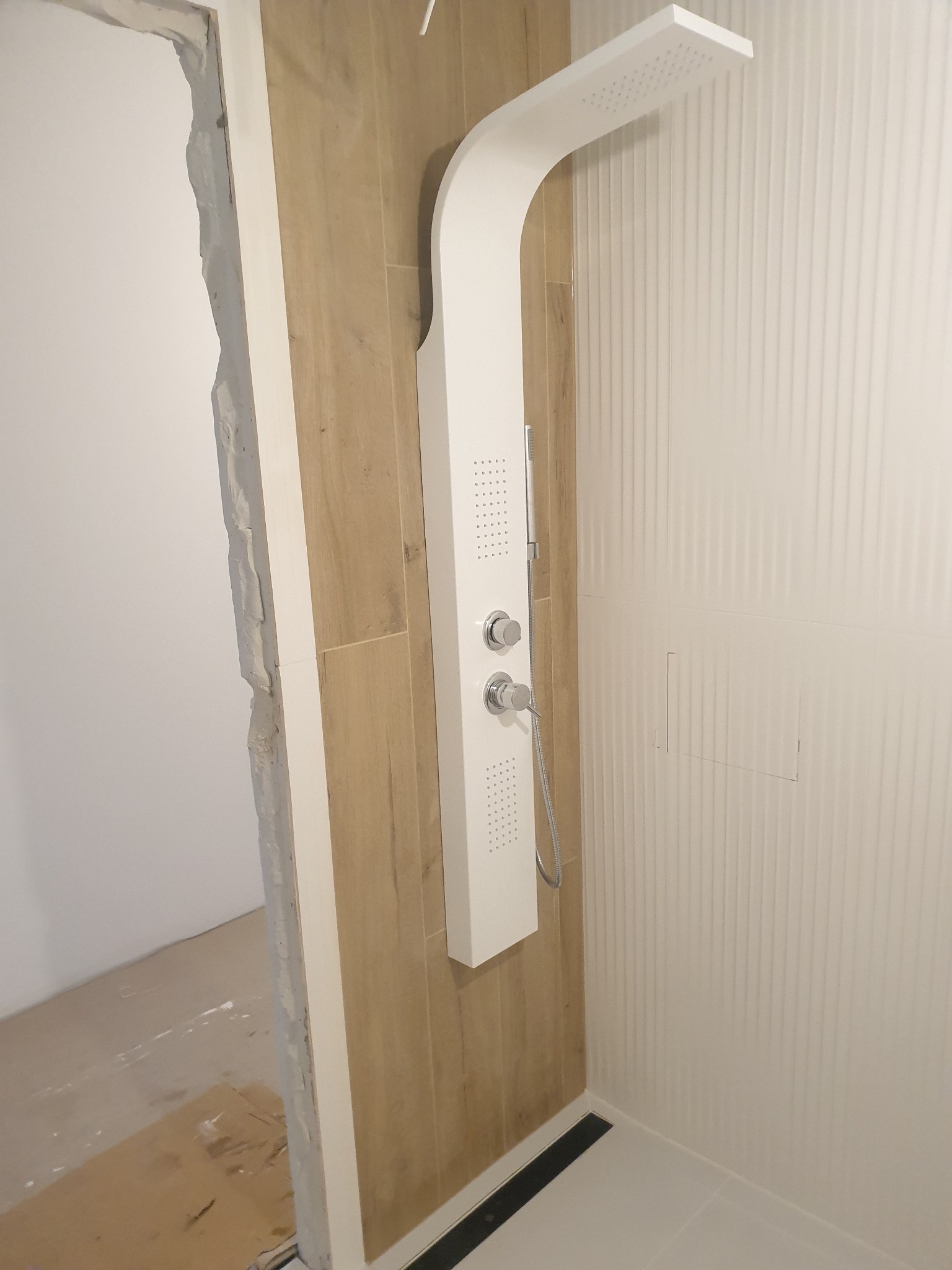 Biały panel prysznicowy z deszczownicą i chromowanymi pokrętłami, zamontowany na ścianie wyłożonej pionowymi, białymi płytkami; obok fragment ściany wyłożonej płytkami imitującymi drewno i surowy...