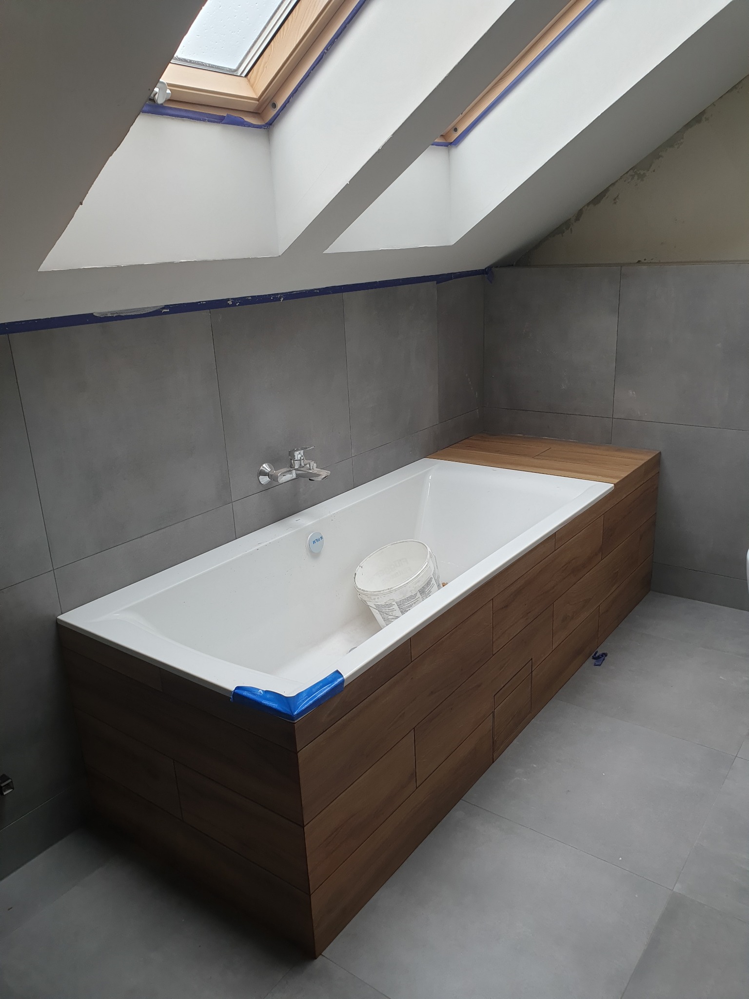 Biała wanna obudowana panelami drewnopodobnymi, z szarą glazurą na ścianach i podłodze, pod skośnym sufitem z oknem dachowym, zabezpieczona niebieską taśmą malarską.