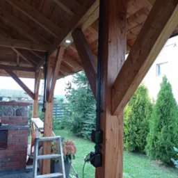 Drewniana altana z widoczną instalacją elektryczną w postaci czarnej rury z gniazdkami, dodatkowo oświetlenie punktowe w suficie, na pierwszym planie drabina i narzędzia.