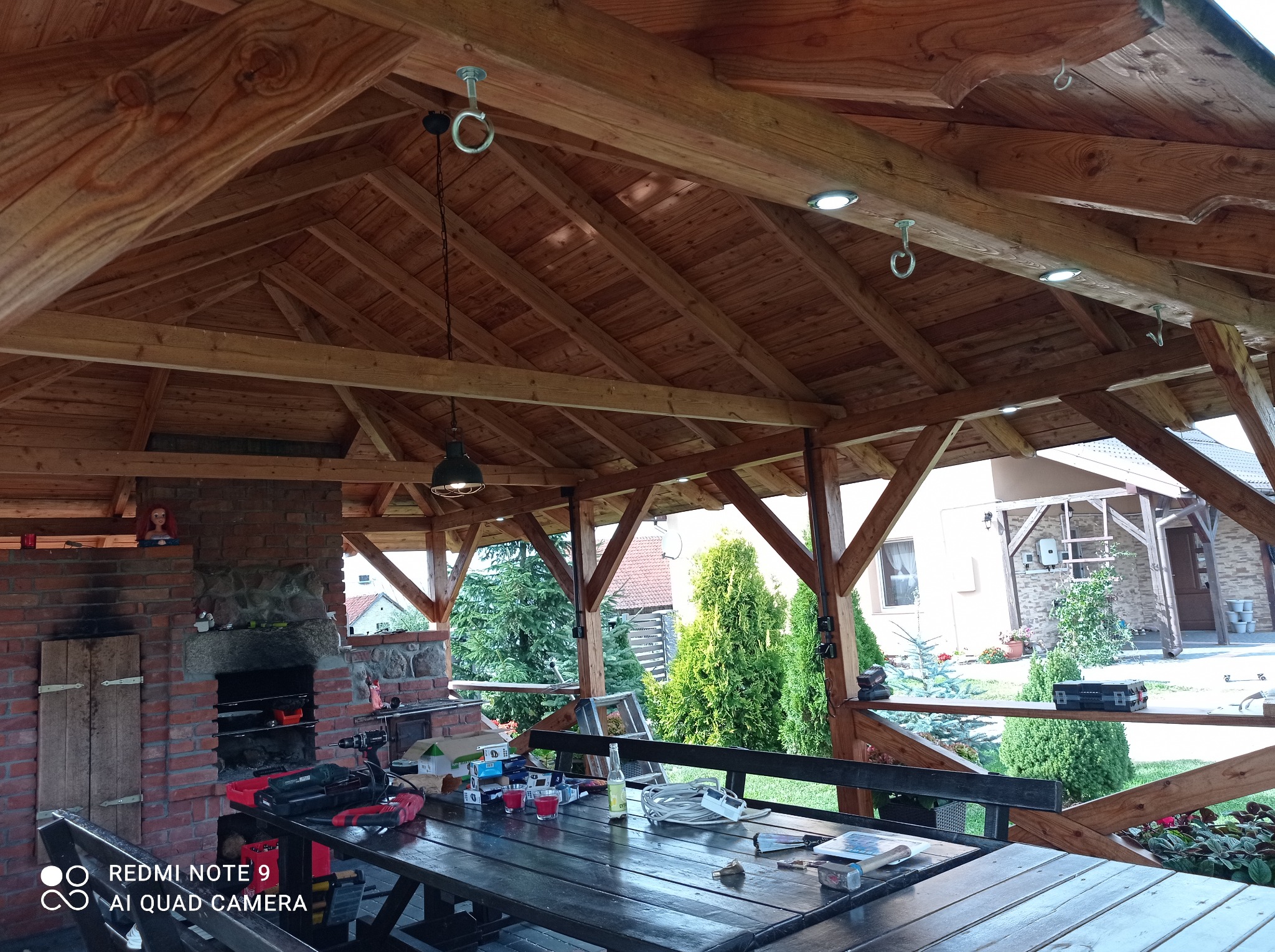 Drewniana altana z oświetleniem punktowym w dachu, murowanym grillem i stołem roboczym z narzędziami, widok na ogród.