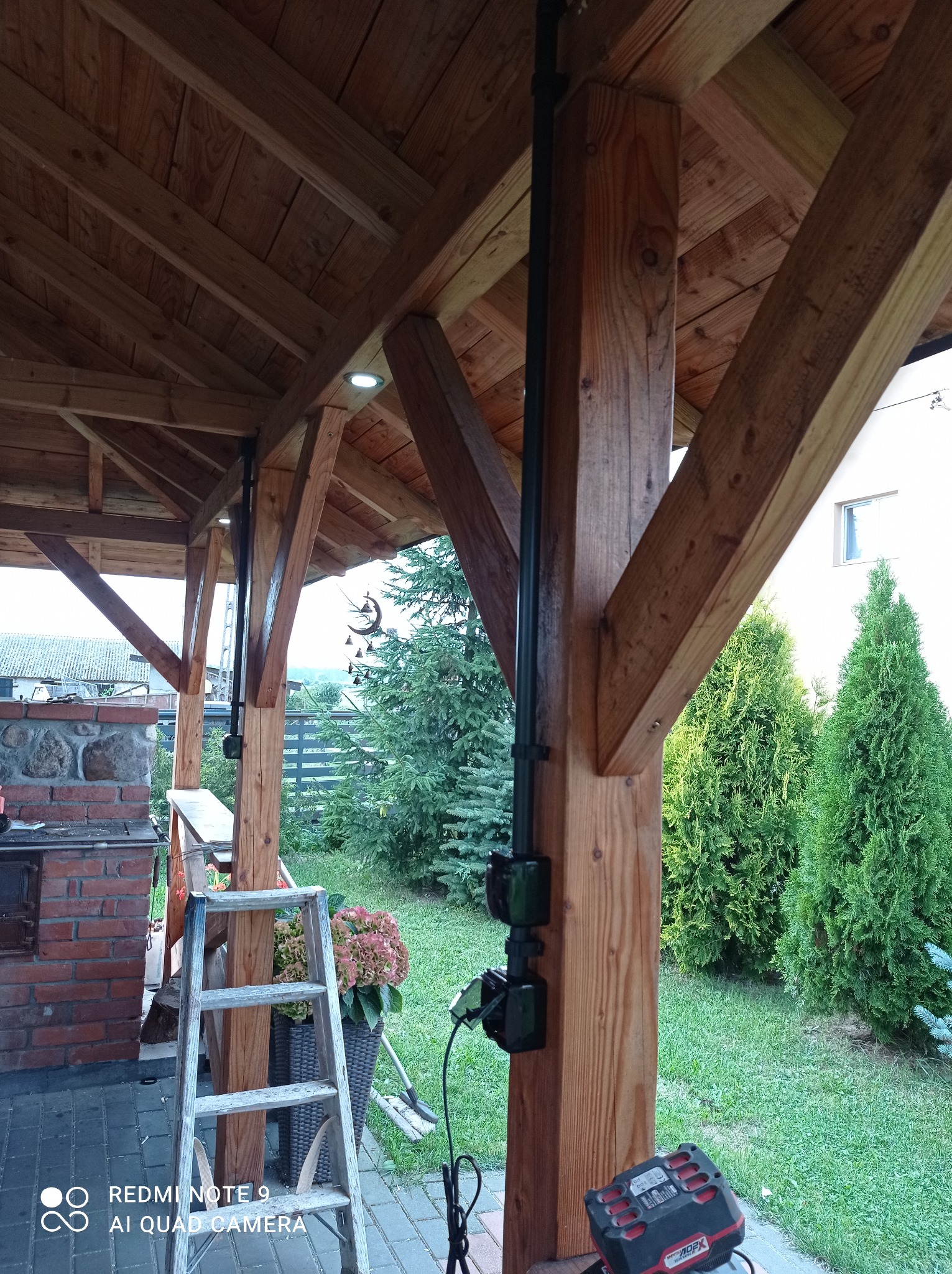 Drewniana altana z widoczną instalacją elektryczną w postaci czarnej rury z gniazdkami, dodatkowo oświetlenie punktowe w suficie, na pierwszym planie drabina i narzędzia.