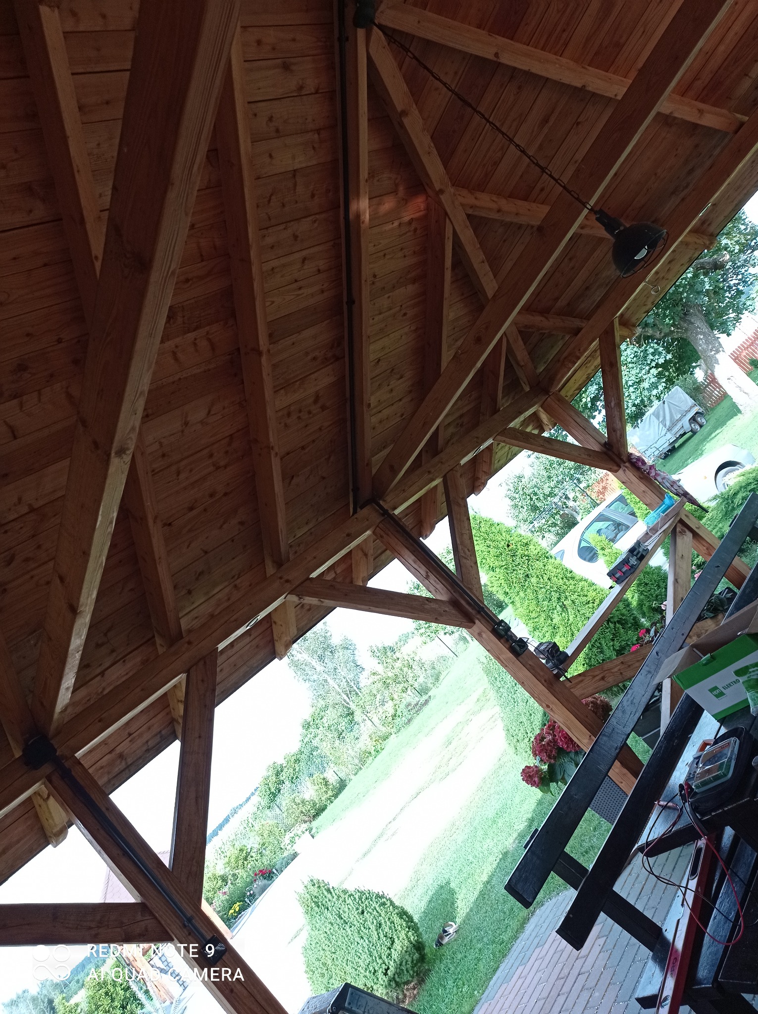 Drewniana konstrukcja altany z widocznym oświetleniem zewnętrznym i narzędziami elektrycznymi na stole, skierowana w stronę ogrodu.