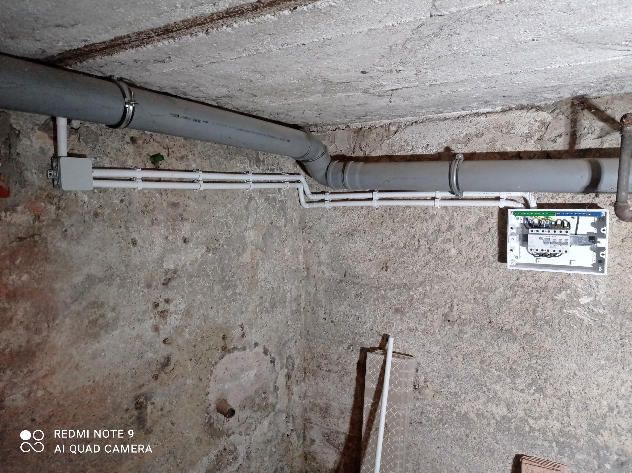 Szare rury PCV przymocowane do sufitu piwnicy za pomocą metalowych obejm, połączone z białą puszką instalacyjną i rozdzielnicą elektryczną na ścianie z surowego betonu.