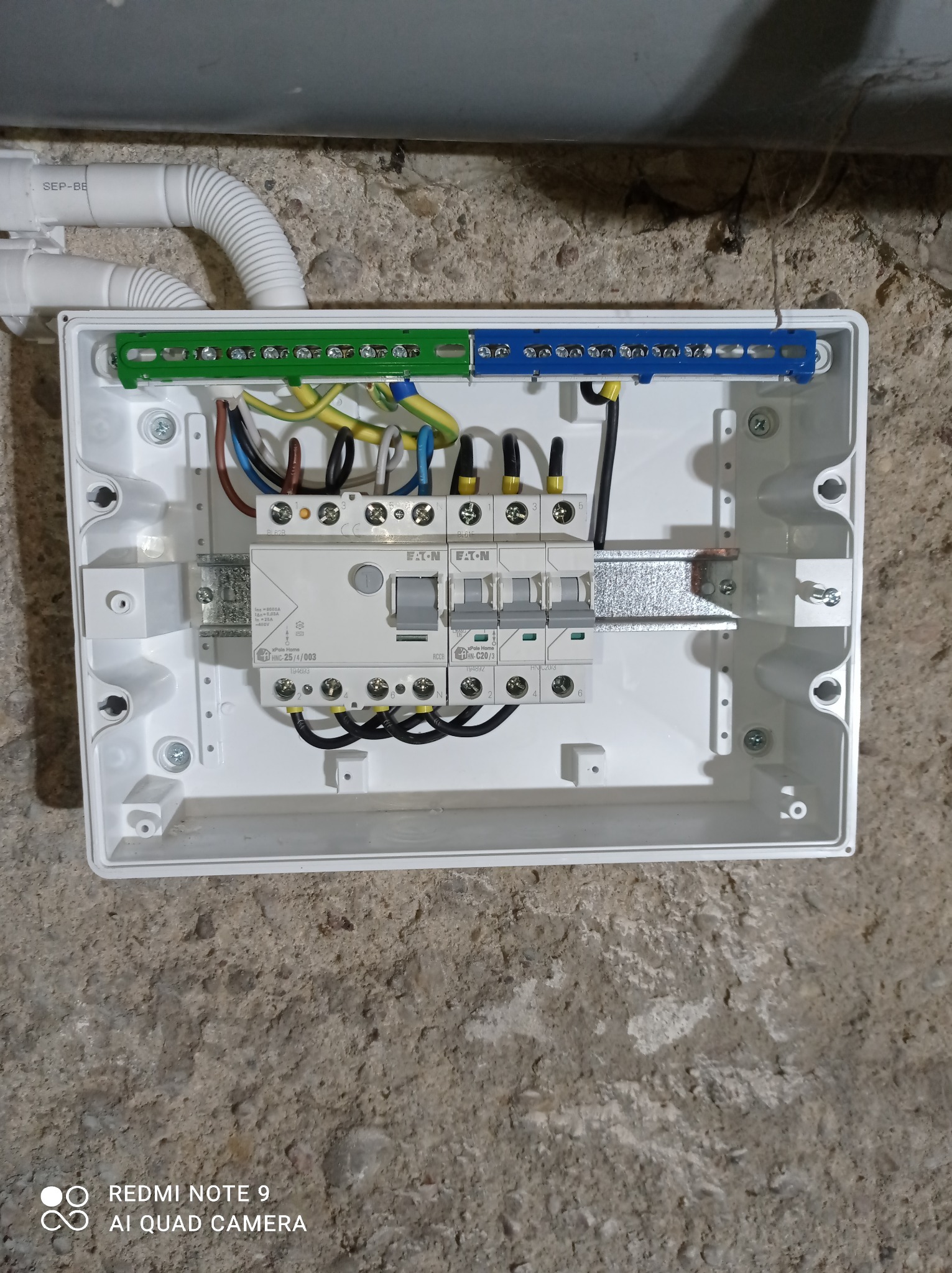 Otwarta skrzynka elektryczna z zainstalowanymi bezpiecznikami Eaton, widoczne przewody w różnych kolorach i listwy zaciskowe, zamontowana na szorstkiej, betonowej ścianie.