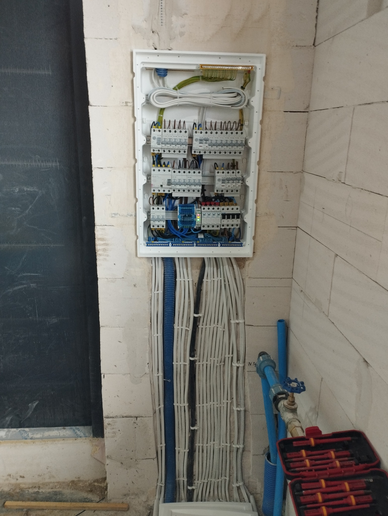 Rozdzielnica elektryczna zamontowana w surowej ścianie z betonu komórkowego, widoczne liczne przewody w peszlach, skrzynka z narzędziami w prawym dolnym rogu.