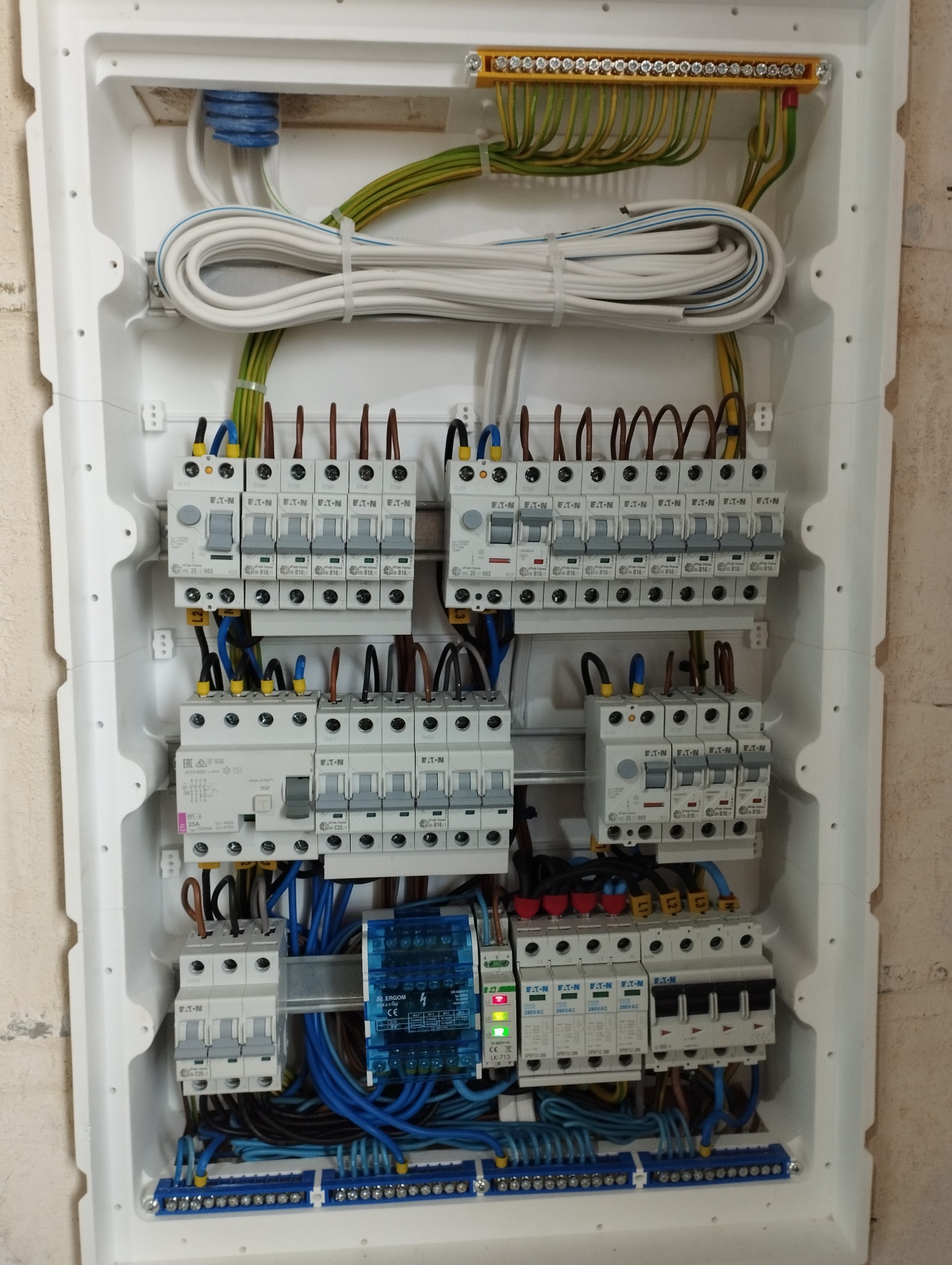 Rozdzielnica elektryczna w białej obudowie, widoczne liczne bezpieczniki Eaton, przewody w różnych kolorach (niebieski, brązowy, żółto-zielony), listwy zaciskowe i moduł ERGOM z diodami...