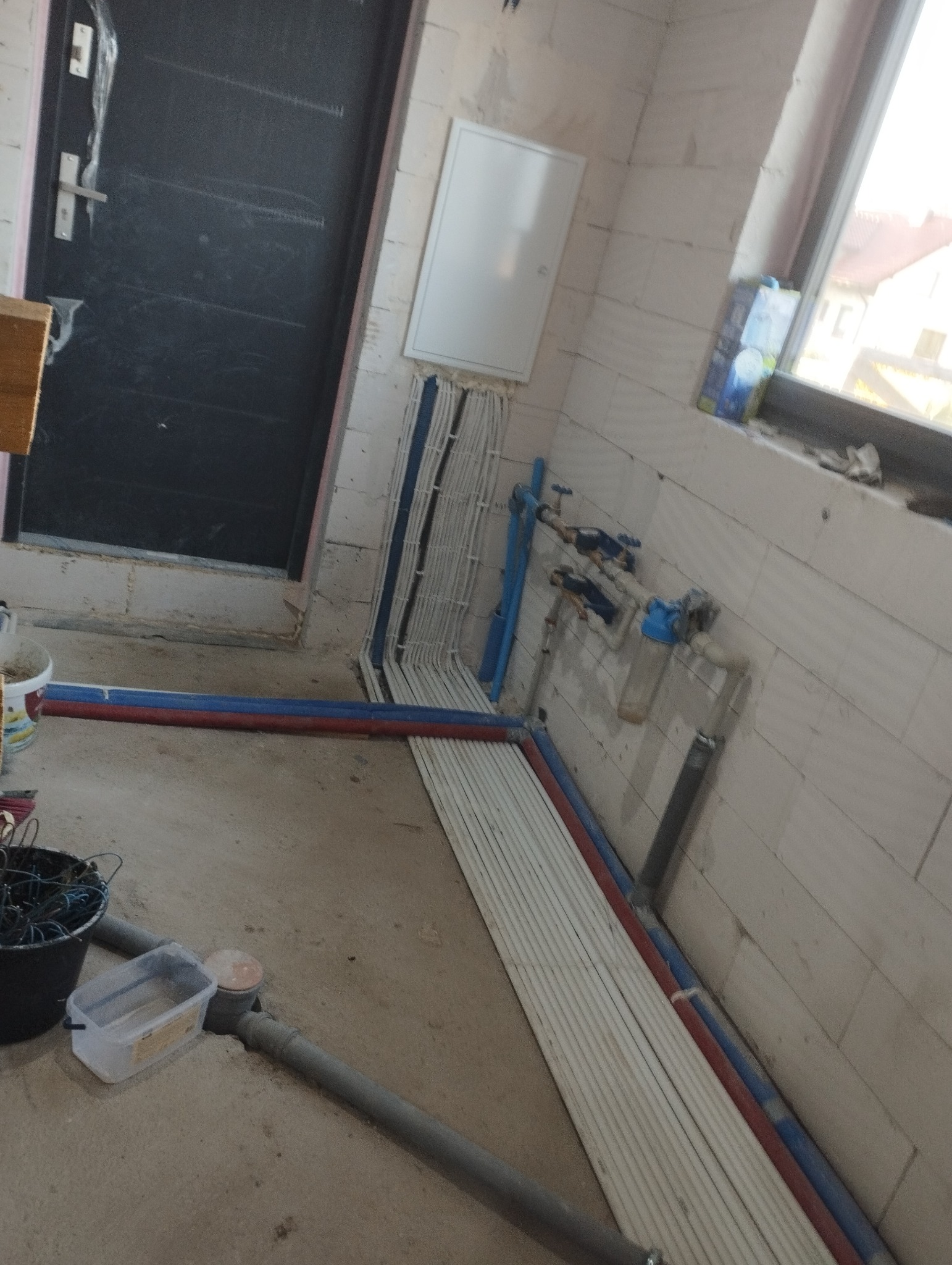 Wnętrze w trakcie prac instalacyjnych: widoczne ułożone kable elektryczne w peszlach, rury wodne, skrzynka rozdzielcza na ścianie z bloków oraz wiadro z przewodami w tle.