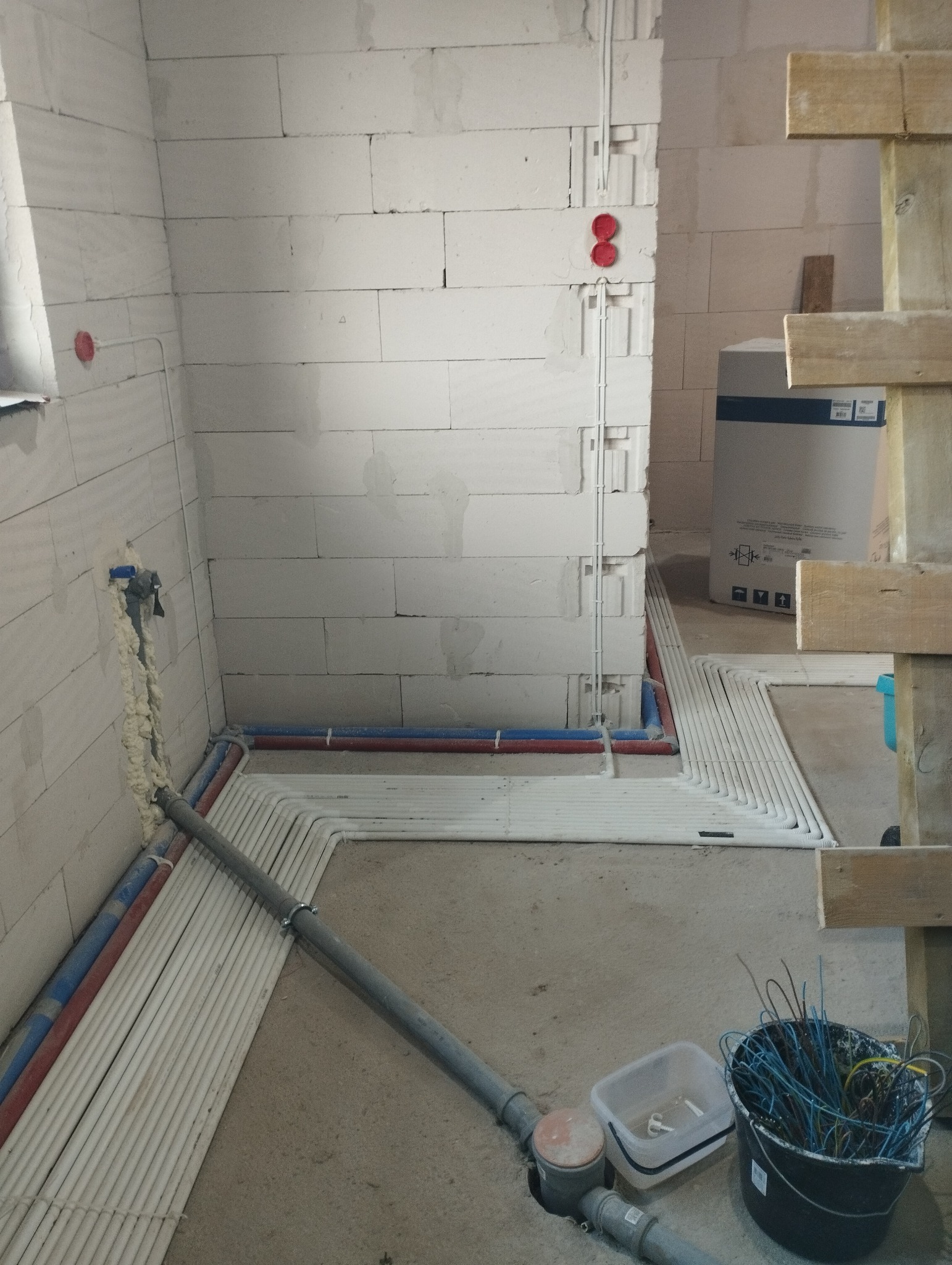 Surowy stan instalacji w pomieszczeniu: przewody elektryczne w białych rurkach ułożone wzdłuż ścian, rury wodne czerwone i niebieskie, puszki instalacyjne w ścianie z czerwonymi zaślepkami, wiadro...