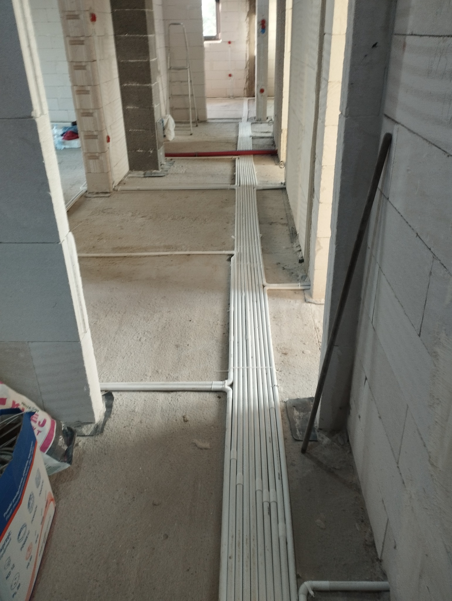Wiązka białych rurek instalacyjnych ułożona równolegle na surowym podłożu wewnątrz budynku w trakcie budowy, widoczne ściany z betonu komórkowego i otwory drzwiowe, drabina w tle.