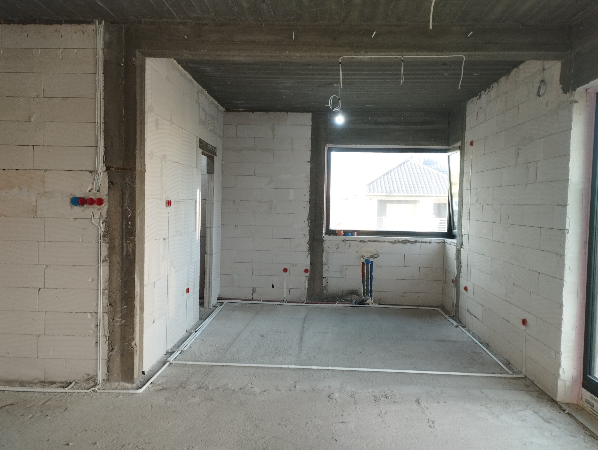 Surowy stan wnętrza: ściany z białego betonu komórkowego, widoczne rury instalacji elektrycznej i wodnej, okno z widokiem na dom z szarą dachówką.