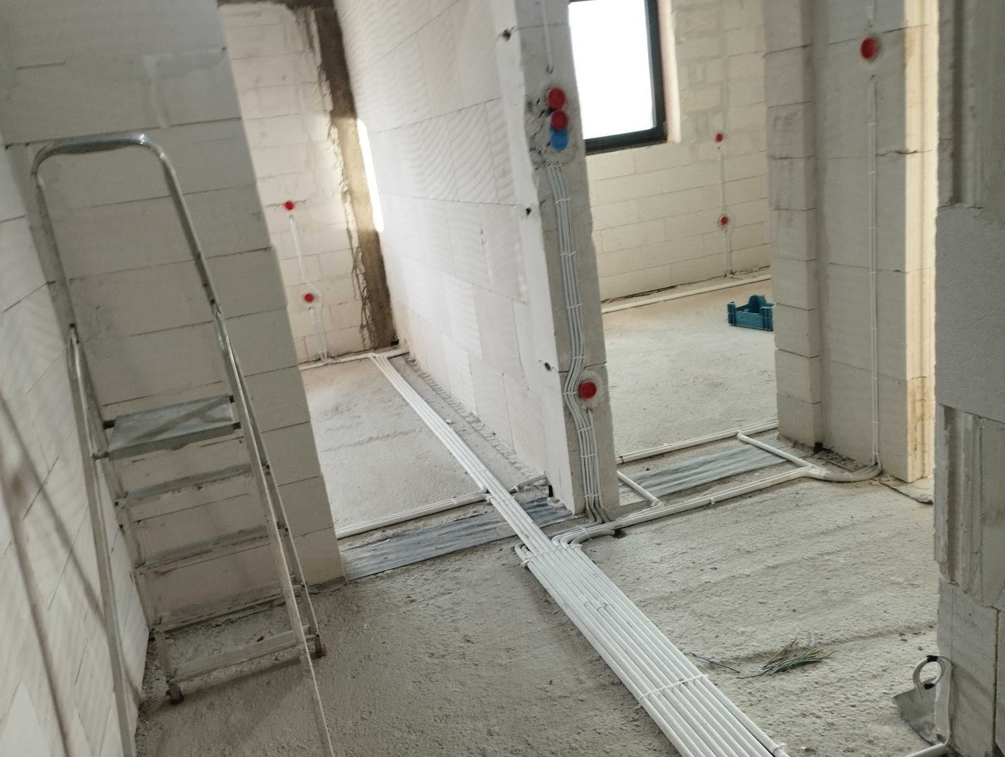 Wnętrze w trakcie instalacji elektrycznej: widoczne białe kable prowadzone po podłodze i ścianach z zamontowanymi puszkami elektrycznymi, drabina oparta o ścianę z betonu komórkowego.