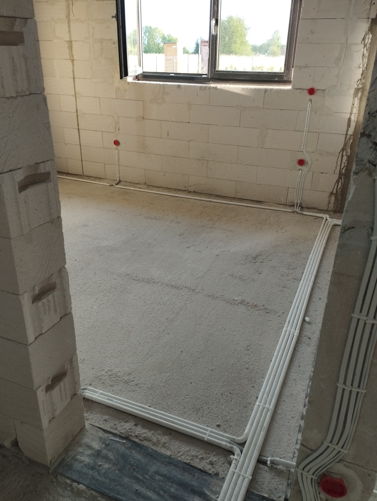 Surowy stan wnętrza w trakcie układania peszli elektrycznych na ścianach z betonu komórkowego i podłodze, widoczne puszki instalacyjne w kolorze czerwonym.