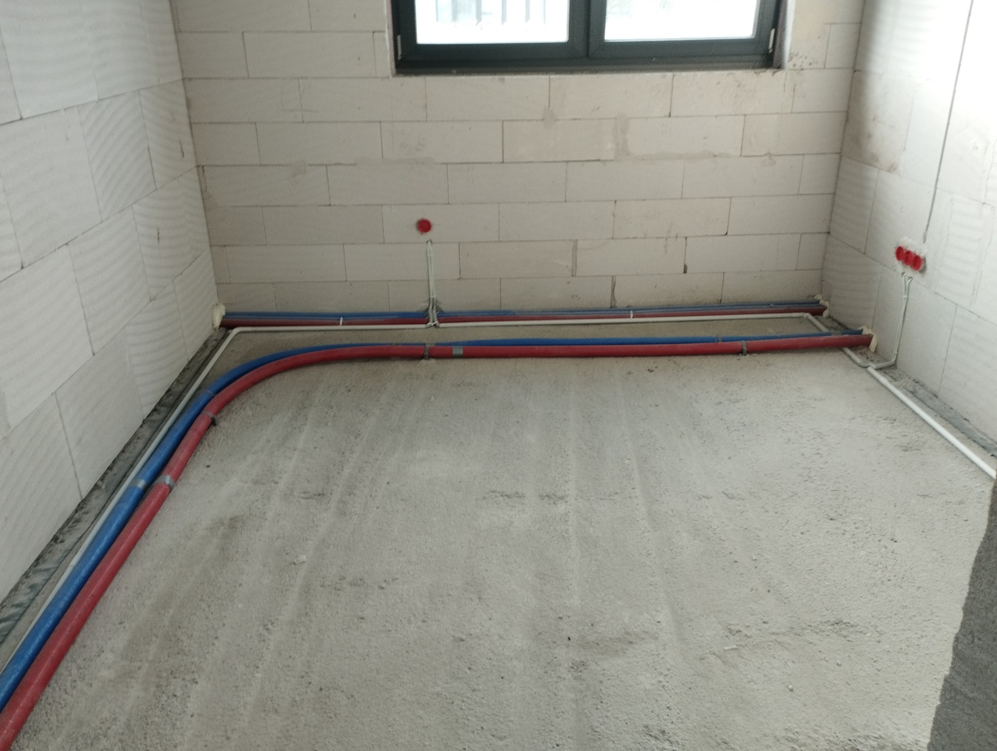 Surowy stan pomieszczenia z widoczną instalacją wodną (rury czerwone i niebieskie) oraz elektryczną (białe kable) poprowadzoną wzdłuż ścian z bloczków betonowych, przed wylewką podłogi.