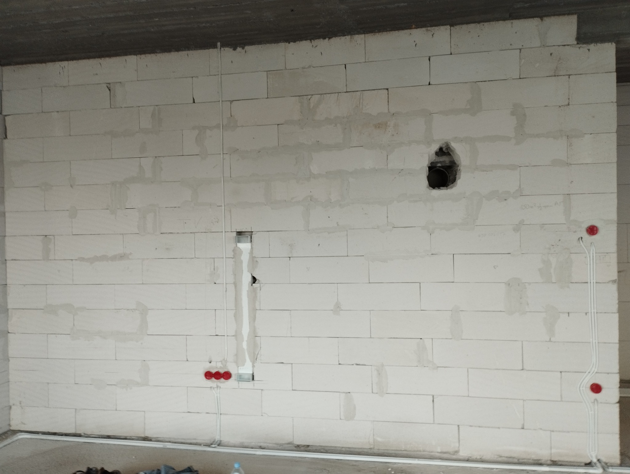 Surowa ściana z betonu komórkowego z widocznymi przewodami elektrycznymi w peszlach, puszkami podtynkowymi w kolorze czerwonym i otworem na rurę kanalizacyjną, etap prac instalacyjnych.