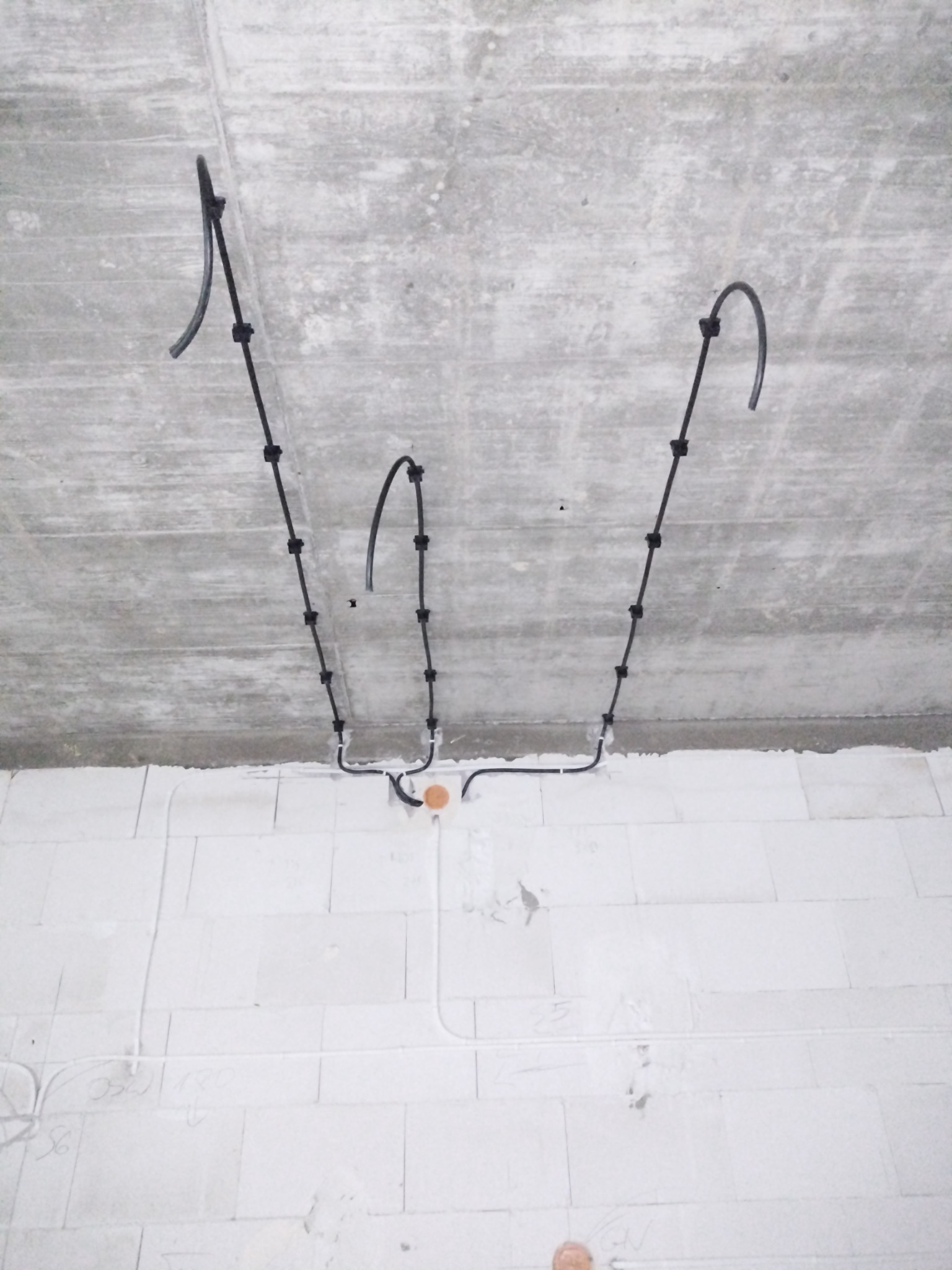 Surowy betonowy strop z trzema czarnymi przewodami elektrycznymi mocowanymi uchwytami, schodzącymi na ścianę z białych bloczków, gdzie widać białe rurki instalacyjne i odręczne notatki.