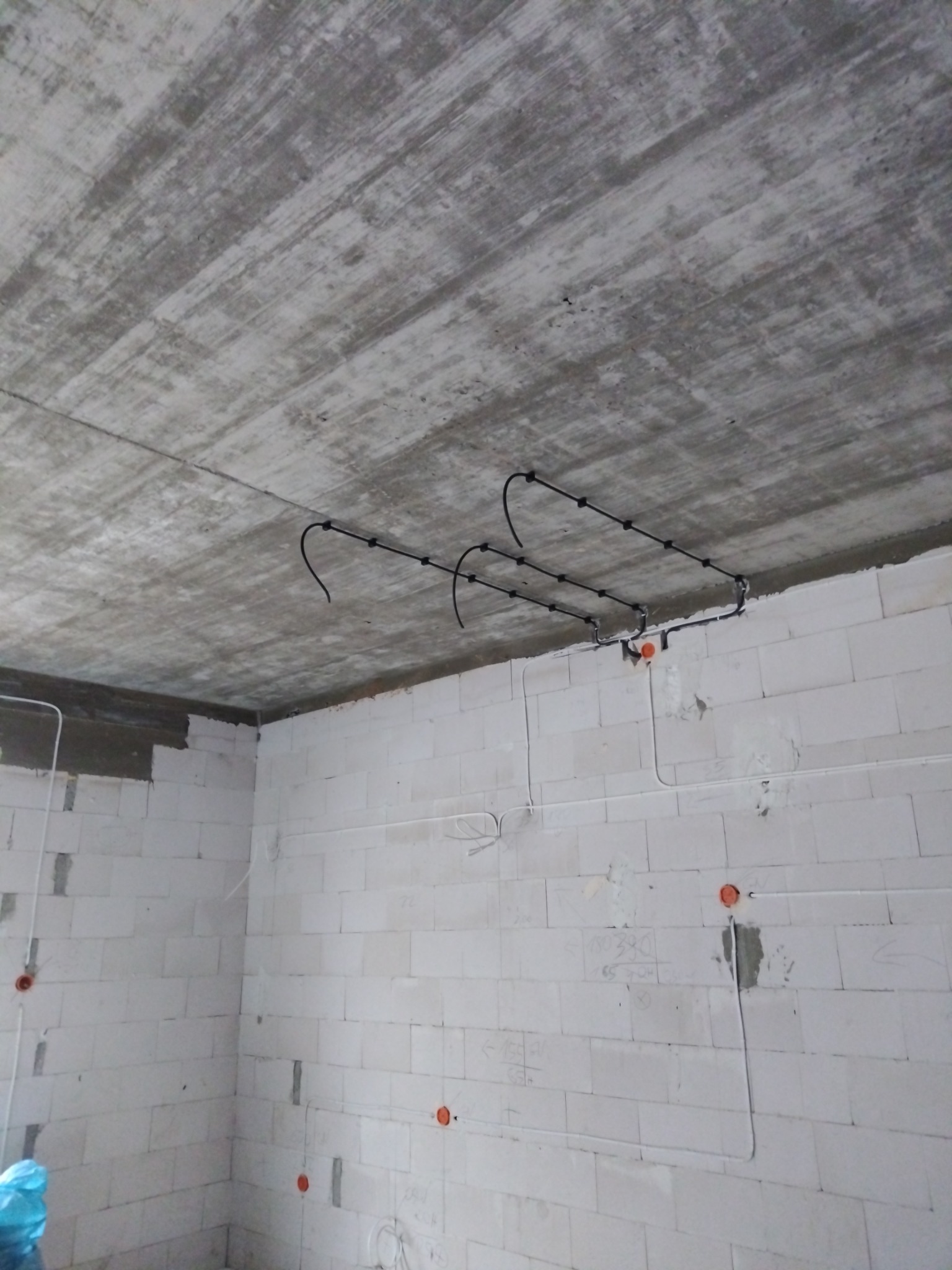 Surowy betonowy strop z podwieszonymi czarnymi rurkami karbowanymi do przewodów elektrycznych, ściany z białych bloczków z widocznymi puszkami instalacyjnymi i prowizorycznymi przewodami.