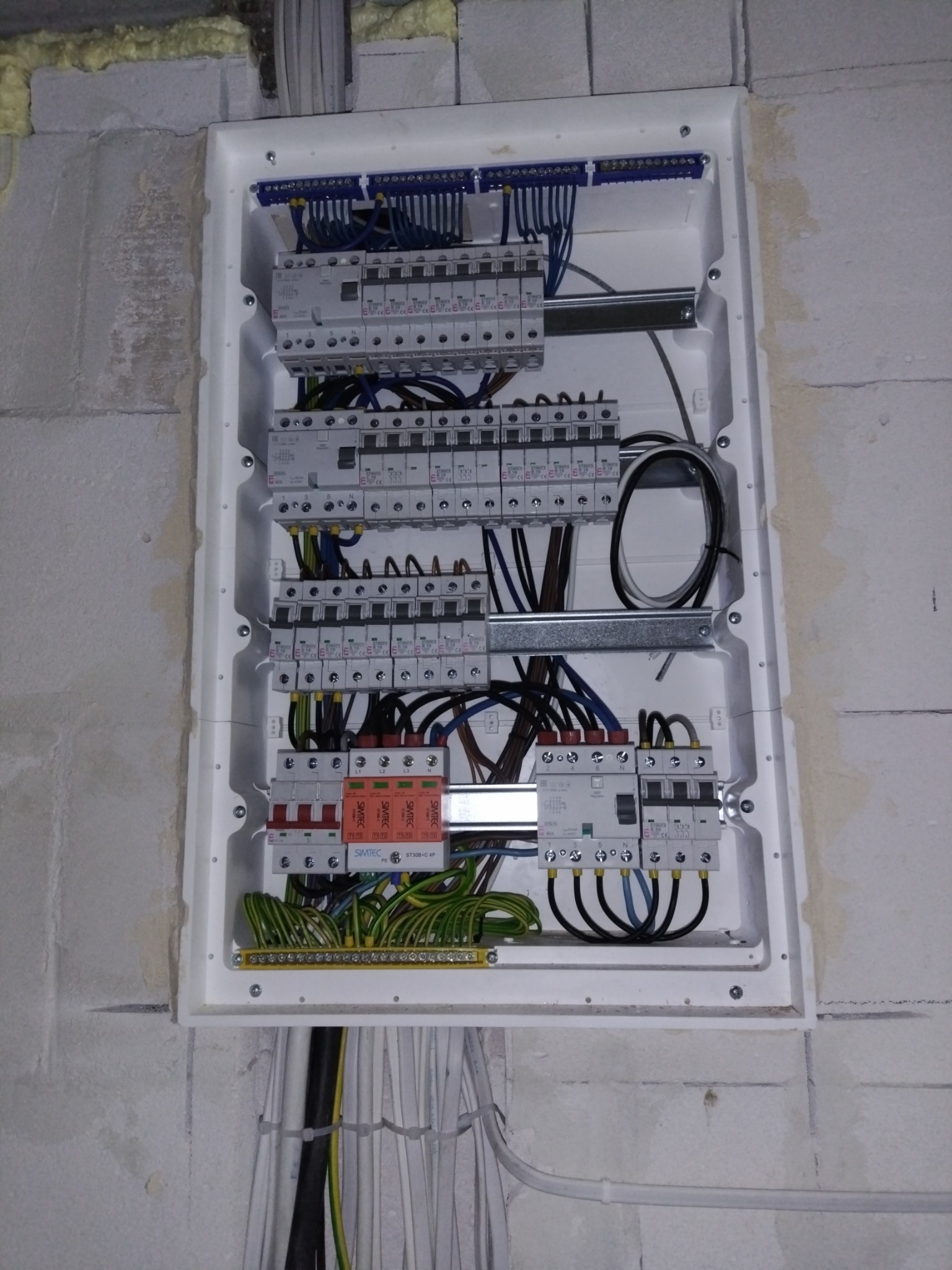 Rozdzielnica elektryczna z zabezpieczeniami różnicowoprądowymi i nadprądowymi, zamontowana na surowej ścianie z betonu komórkowego, widoczne przewody w peszlach wchodzące do skrzynki.