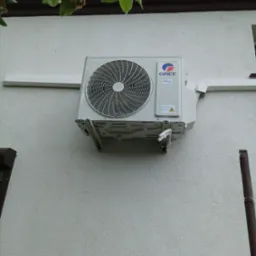 Zewnętrzna jednostka klimatyzatora marki Gree zamontowana na ścianie budynku z widocznymi elementami instalacji odprowadzającej skropliny i fragmentem rynny.