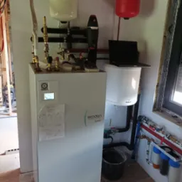 Biała pompa ciepła Ecodan Mitsubishi Electric w trakcie montażu, widoczne rury, zbiorniki wyrównawcze, filtry, laptop na zbiorniku Galmet, podłoga w trakcie wykańczania.