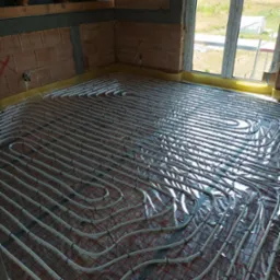 Widok na rozłożone rury ogrzewania podłogowego na folii izolacyjnej w surowym stanie, widoczne ceglane ściany i okno z widokiem na zewnątrz.