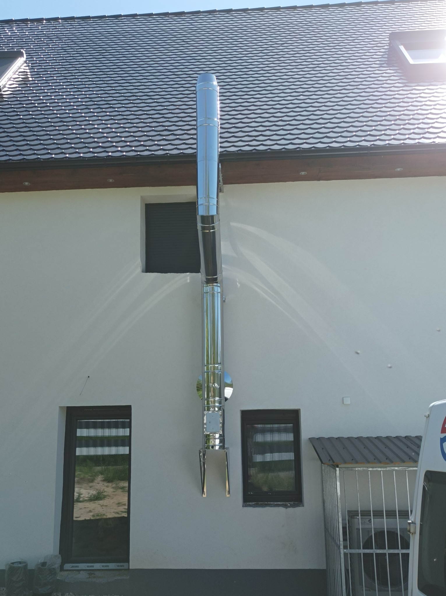 Zewnętrzny, pionowy, metalowy komin spalinowy przymocowany do ściany domu z szarym dachem, widok z dołu na tle błękitnego nieba.