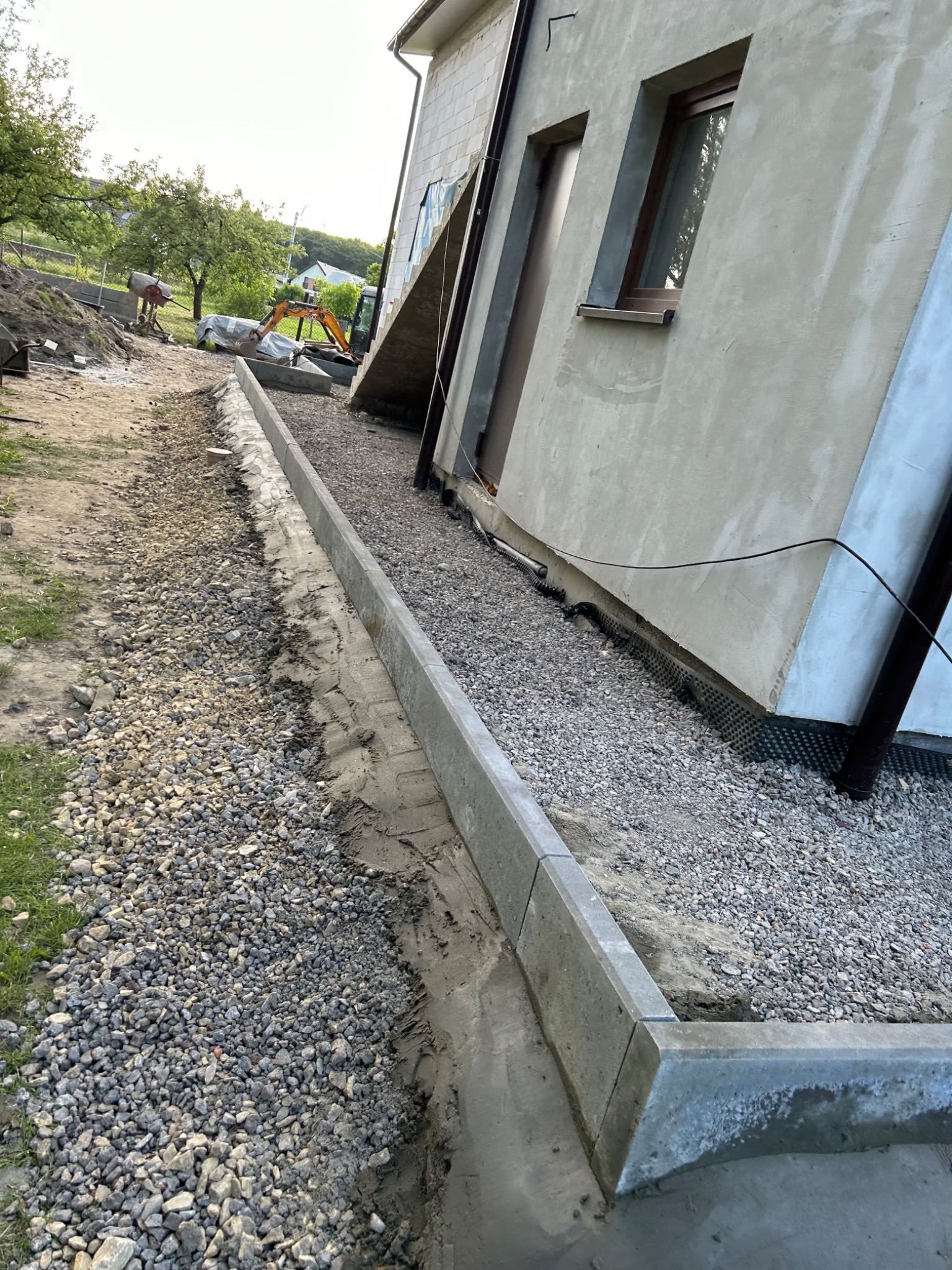 Przygotowanie terenu wokół budynku: ułożone obrzeża betonowe, wysypany żwir, w tle widoczny mały sprzęt budowlany i mieszalnik.