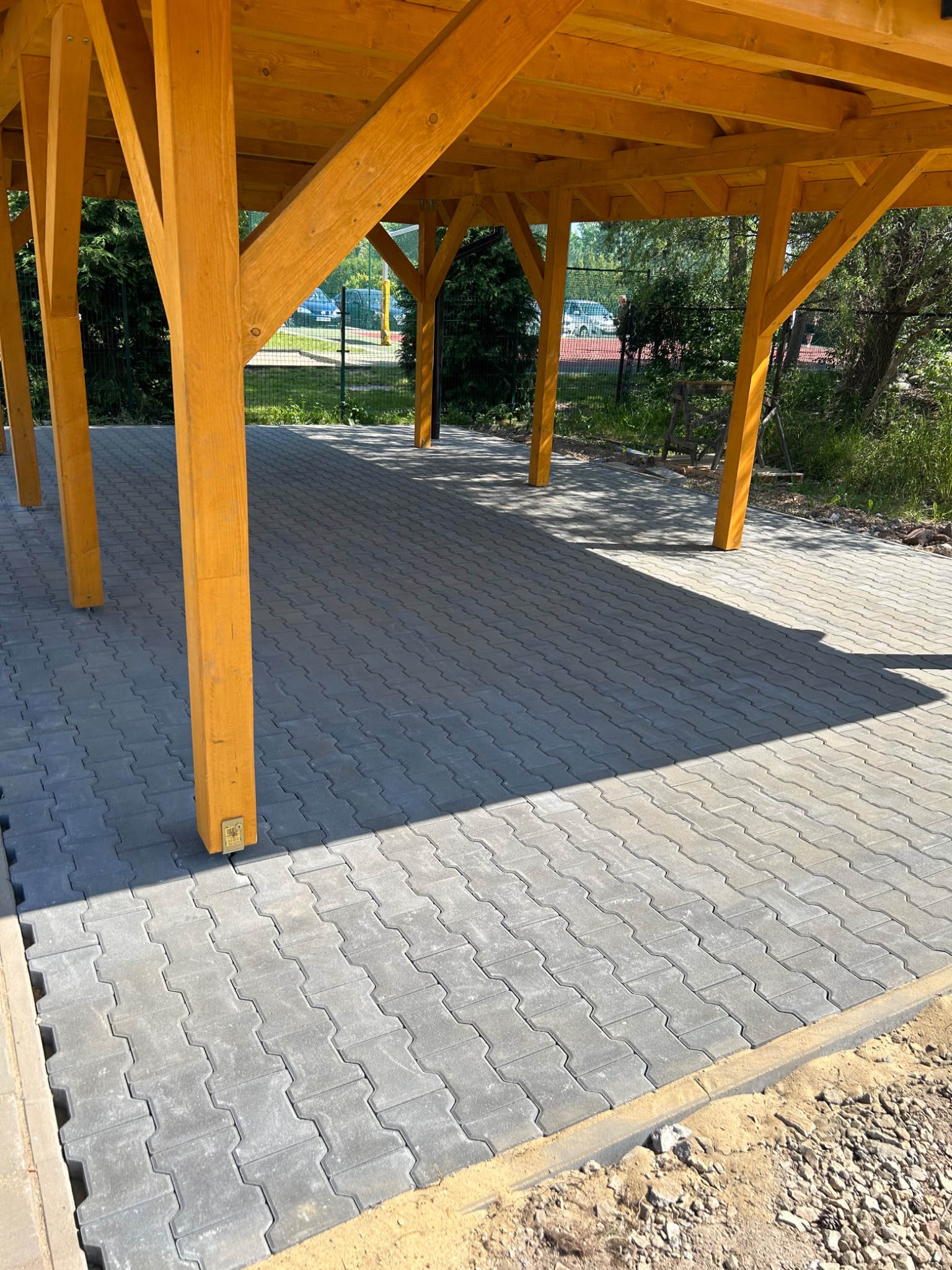 Szara kostka brukowa ułożona pod drewnianą konstrukcją z żółtymi słupami, widoczne krawężniki i teren przygotowany do dalszych prac.
