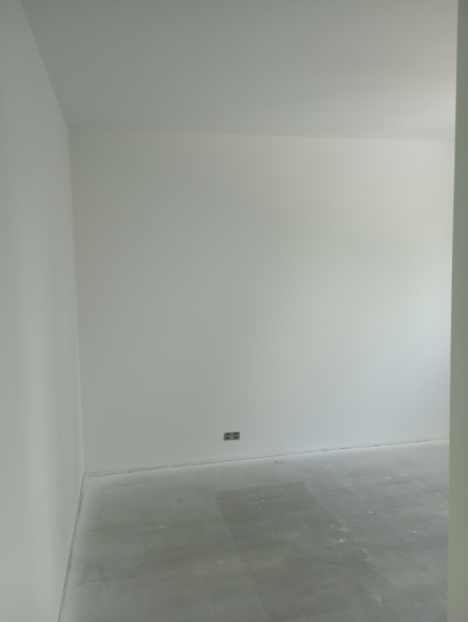 Pomalowane na biało wnętrze pokoju z widocznym gniazdkiem elektrycznym i szarą podłogą w stanie surowym, widok zza framugi drzwi.