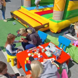 Kobieta maluje wzór na ramieniu dziewczynki siedzącej na żółtym krzesełku przy czerwonym stole, na którym leżą farby i akcesoria do malowania twarzy; w tle dmuchany zamek i bawiące się dzieci.