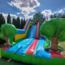 Dzieci zjeżdżają z dmuchanej zjeżdżalni w motywie dżungli, z nadrukowanymi zwierzętami i dekoracjami w kształcie drzew, na tle zielonej trawy i błękitnego nieba z chmurami.