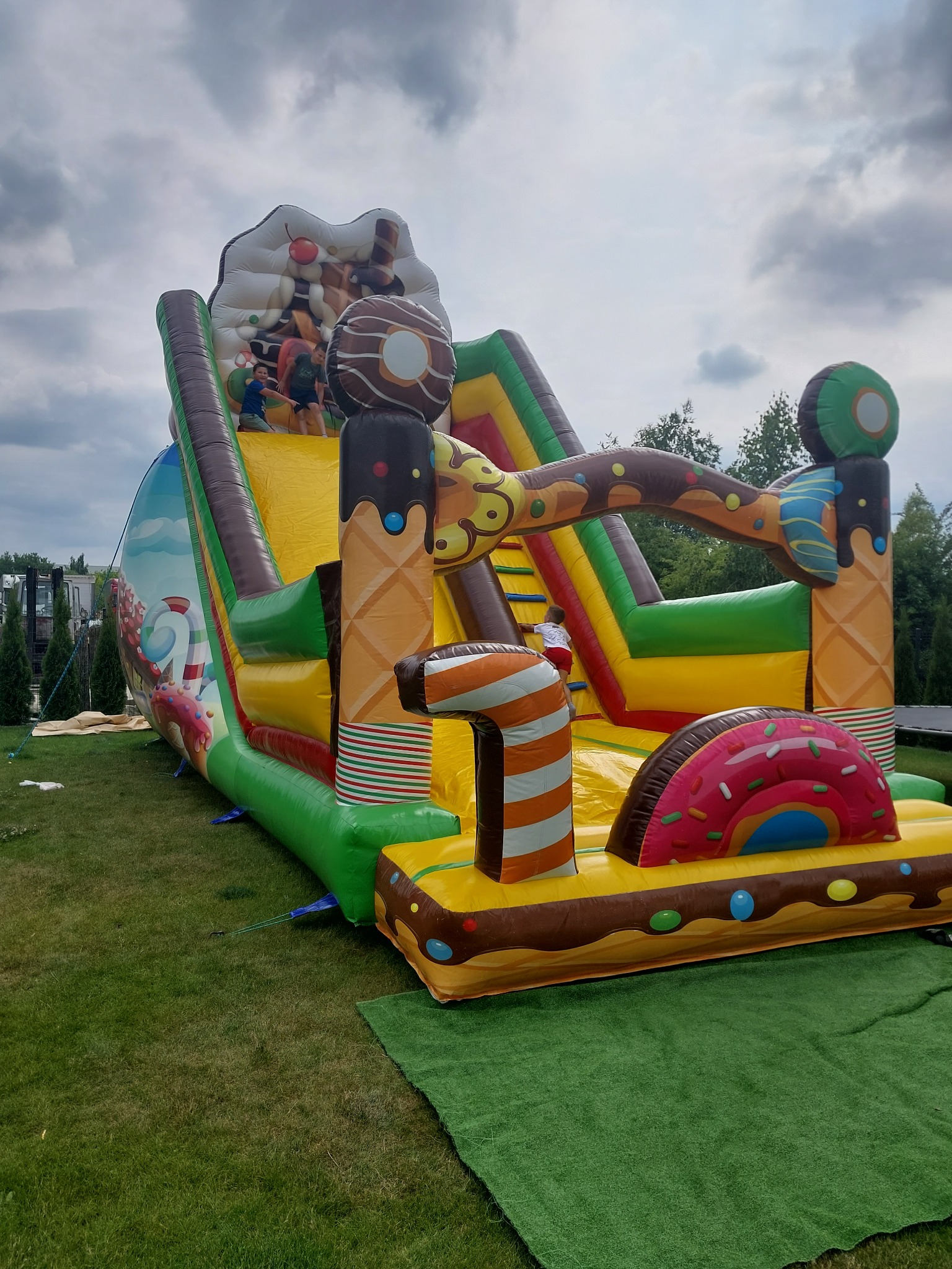 Kolorowy dmuchany plac zabaw z motywem słodyczy, imitujący lody i pączki, ustawiony na trawie, z dziećmi wspinającymi się na górę.