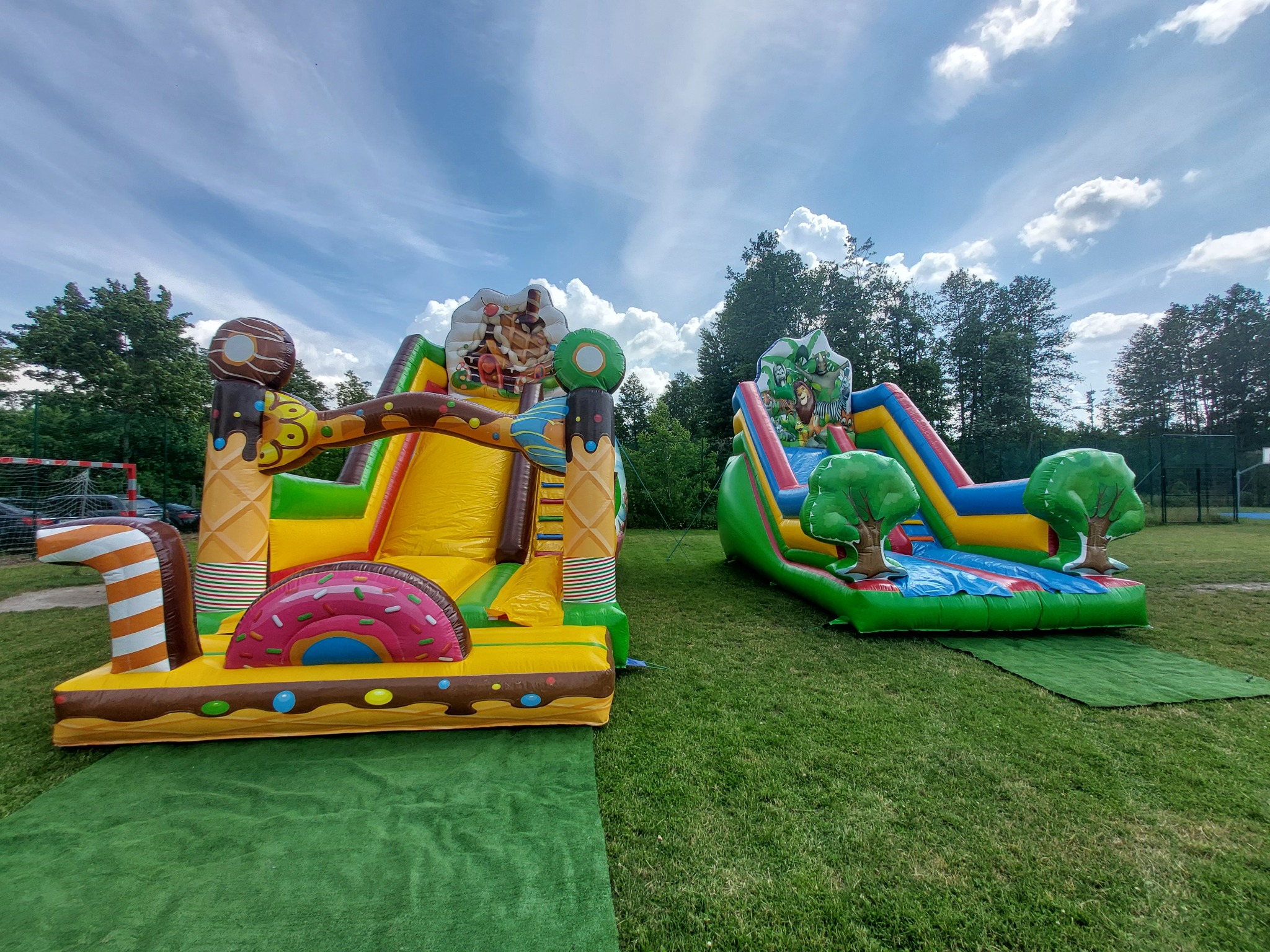 Dwa kolorowe, nadmuchiwane zamki na zielonej trawie: jeden w kształcie lodów i słodyczy, drugi z motywem dżungli i drzew. Na tle błękitne niebo z białymi chmurami.