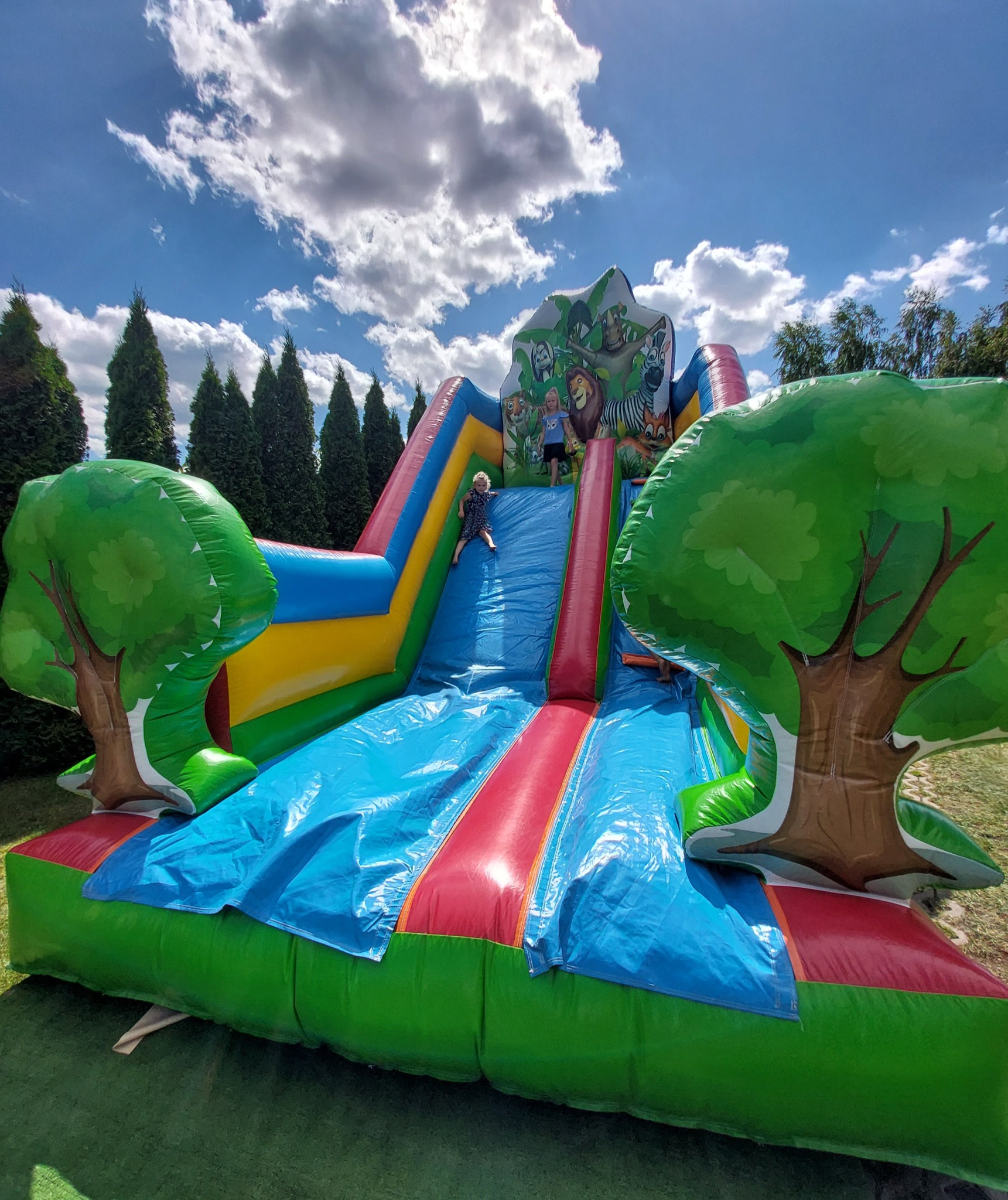 Dzieci zjeżdżają z dmuchanej zjeżdżalni w motywie dżungli, z nadrukowanymi zwierzętami i dekoracjami w kształcie drzew, na tle zielonej trawy i błękitnego nieba z chmurami.