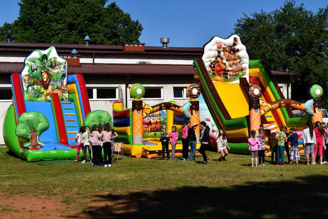Dwa dmuchańce: jeden z motywem dżungli i zjeżdżalnią, drugi przypominający słodycze; dzieci w różnym wieku stoją i bawią się na trawie przed dmuchańcami.