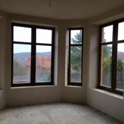 Wnętrze surowego pokoju z trzema oknami z brązowymi ramami, ściany po tynkowaniu, widok na zewnątrz na zabudowę jednorodzinną i drzewa.