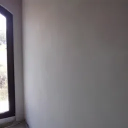 Wnętrze z oknem łukowym i świeżo otynkowaną ścianą w odcieniach szarości, widok na zewnątrz na zieleń.