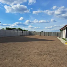 Przygotowany teren pod trawnik z widocznymi śladami po maszynach, ogrodzony betonowym płotem, z geodezyjnym niwelatorem obrotowym na statywie przy budynku, błękitne niebo z chmurami.