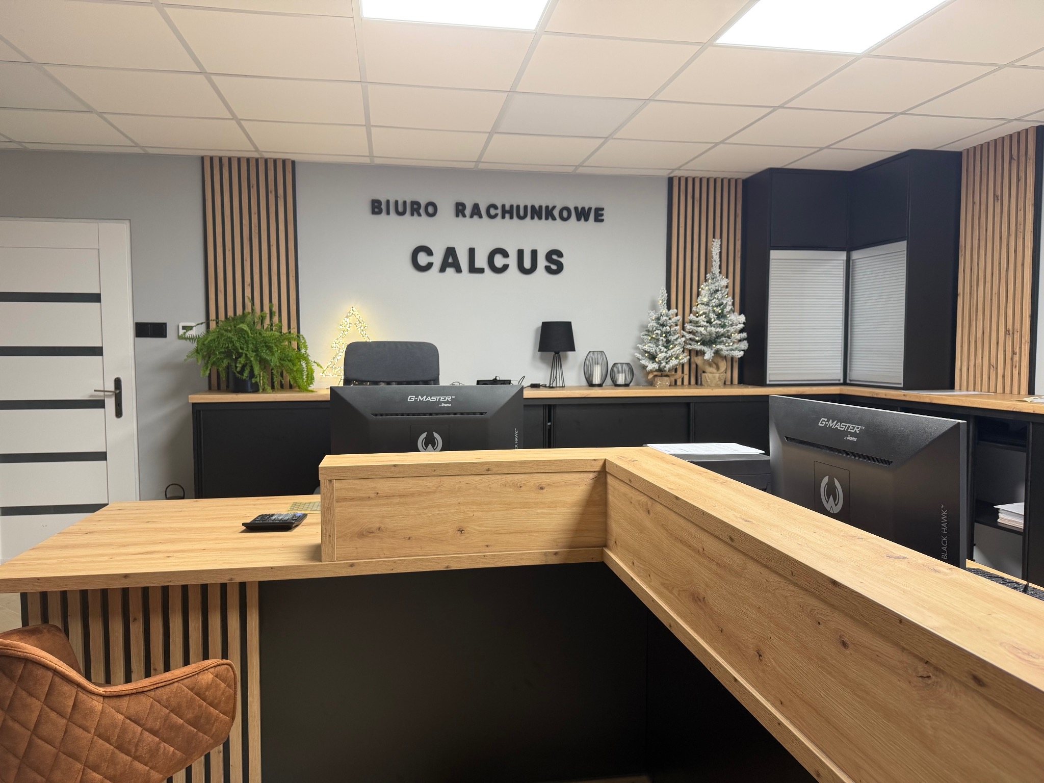 Nowoczesne biuro rachunkowe z drewnianymi akcentami i logo 'Calculus' na ścianie. Na biurku monitory G-Master i dekoracje świąteczne.