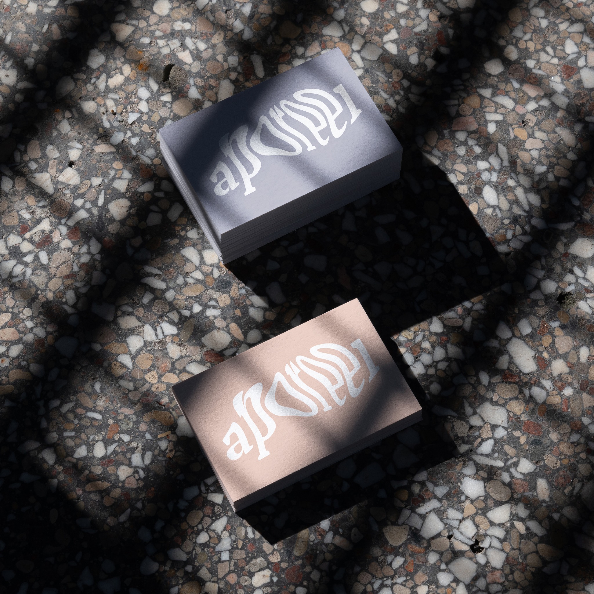 Dwa stosy wizytówek z białym, abstrakcyjnym logo na szarym i brzoskwiniowym tle, rzucające cienie na mozaikową powierzchnię.